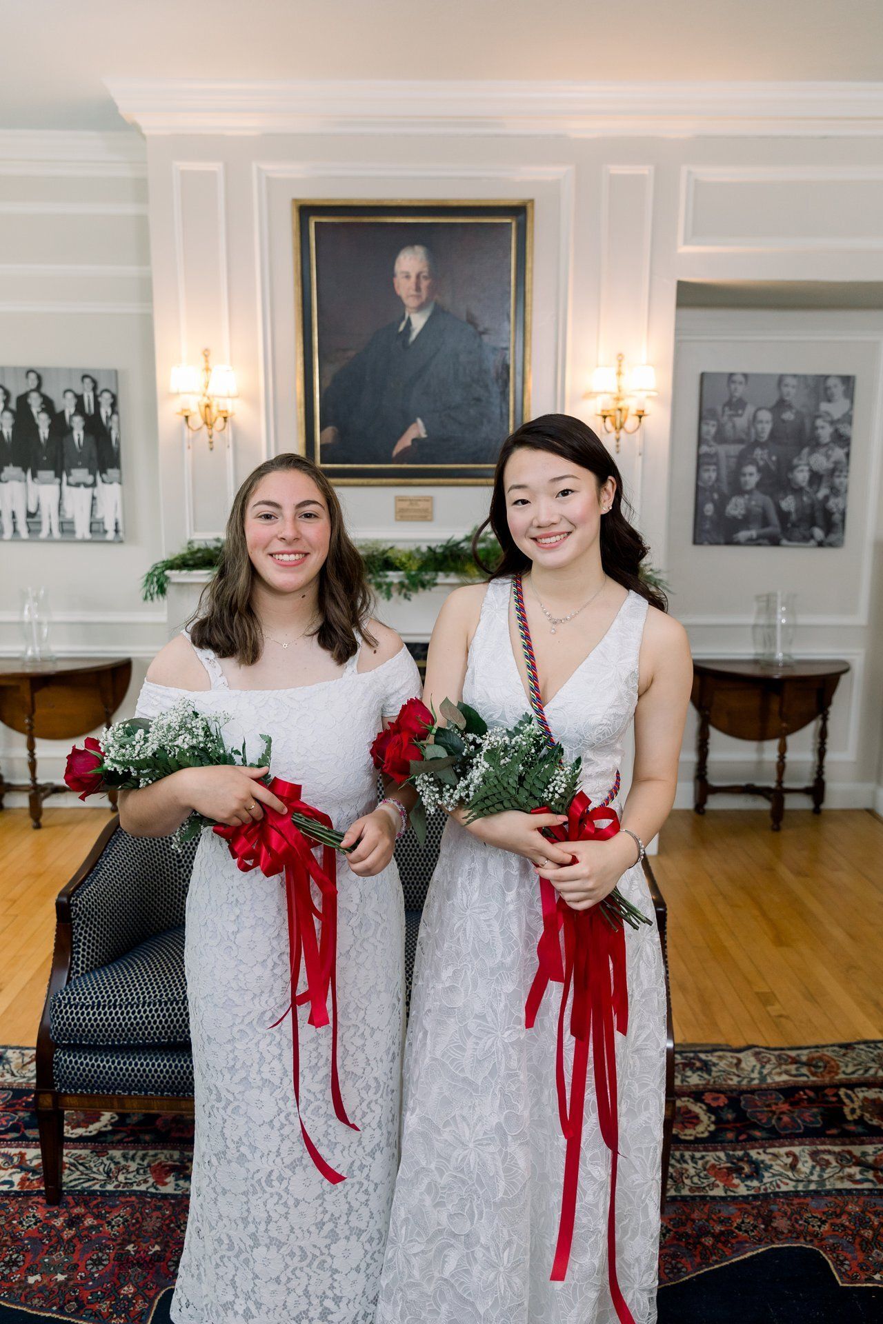 Two women in white dresses holding red-and-white bouquets in a formal room with portraits on the wall