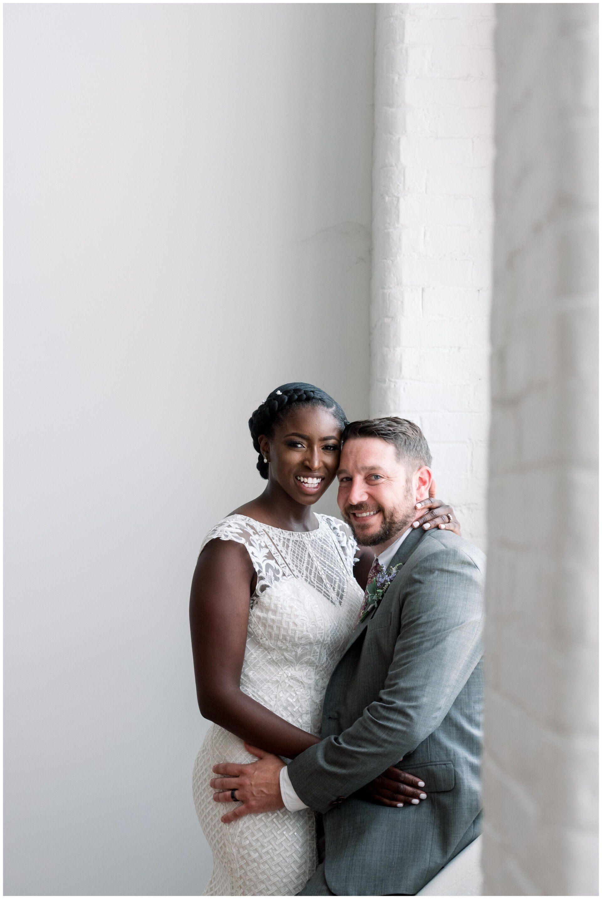 Couple embracing by a bright window, smiling in elegant white and gray attire.