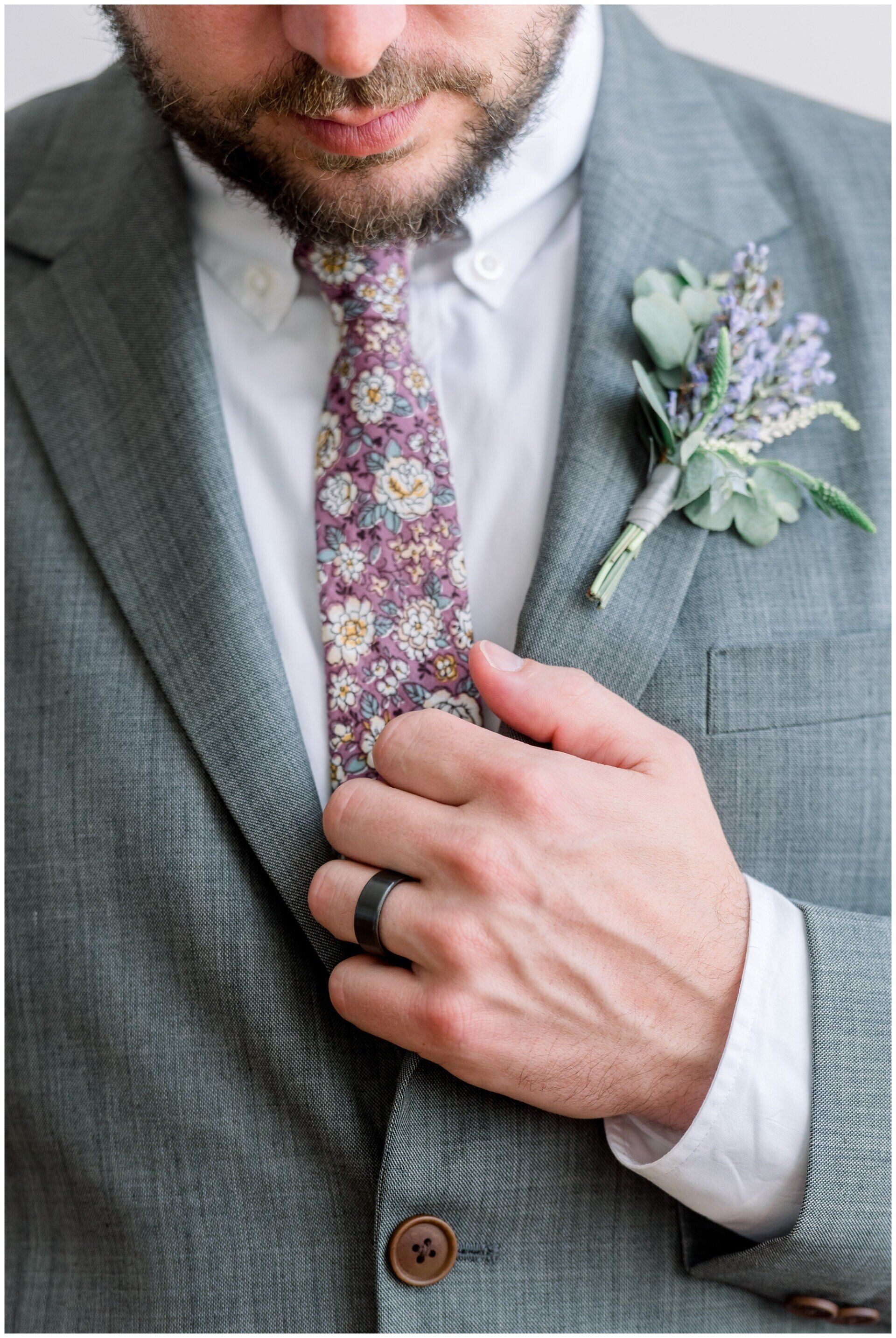 Man adjusting floral tie on gray suit with boutonniere and wedding band