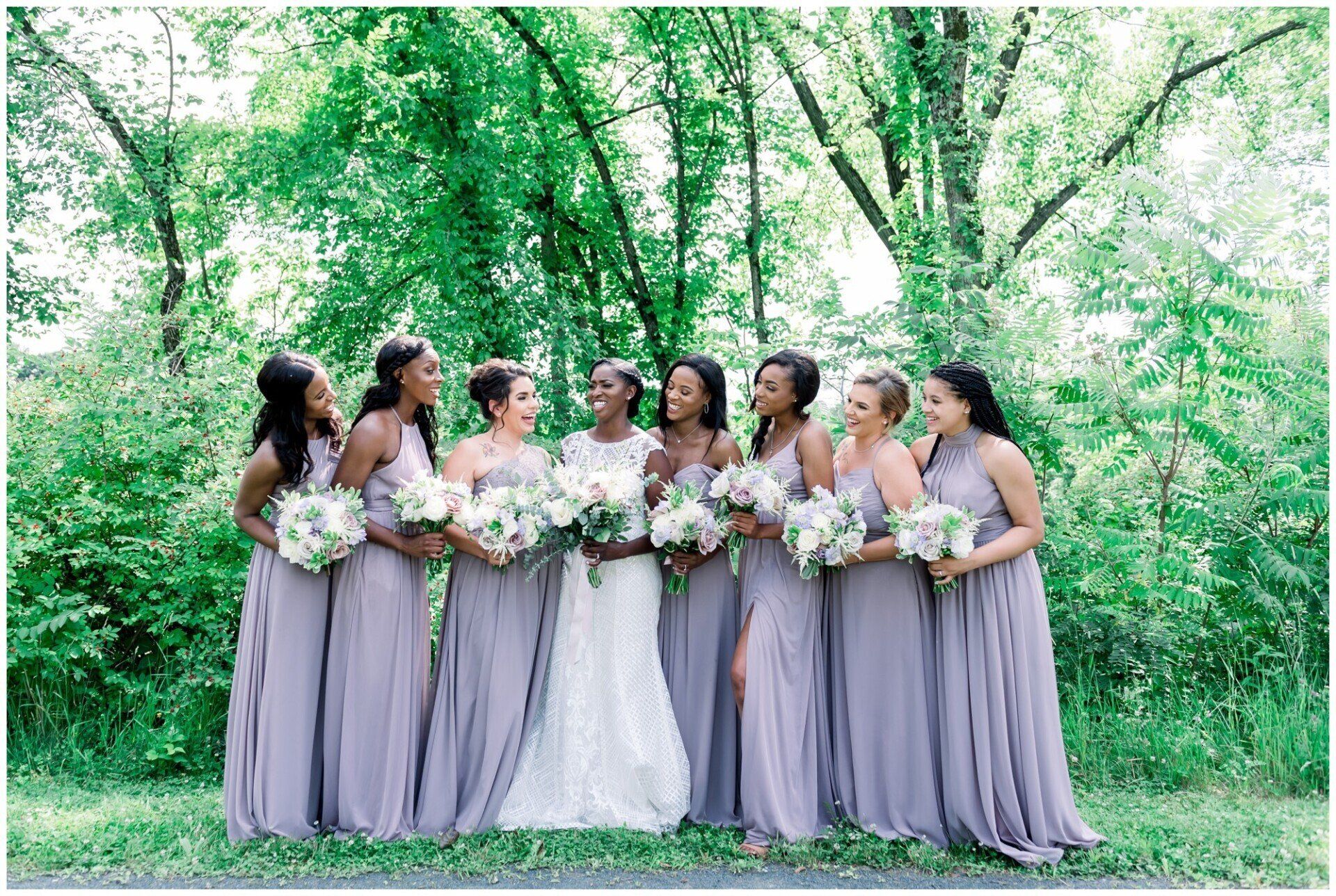 Bridesmaids in lavender dresses holding white bouquets in a green wooded outdoor setting