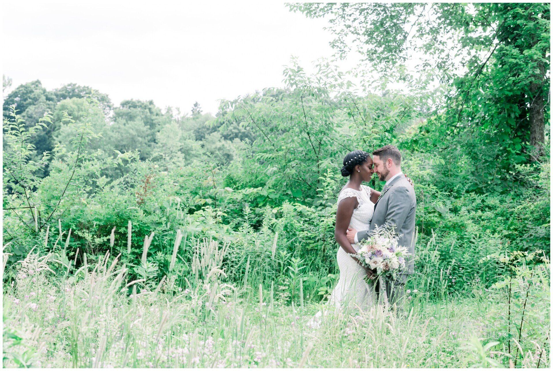 Bride and groom embracing in a lush green meadow, holding a bouquet, under trees.