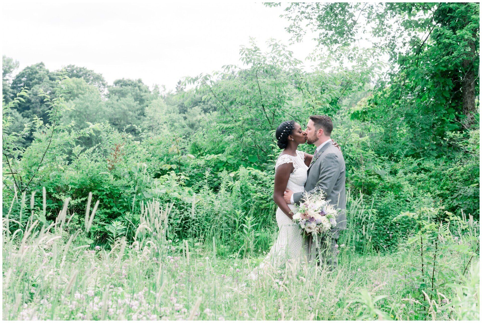 Bride and groom standing in a lush green meadow, facing each other and holding a bouquet.