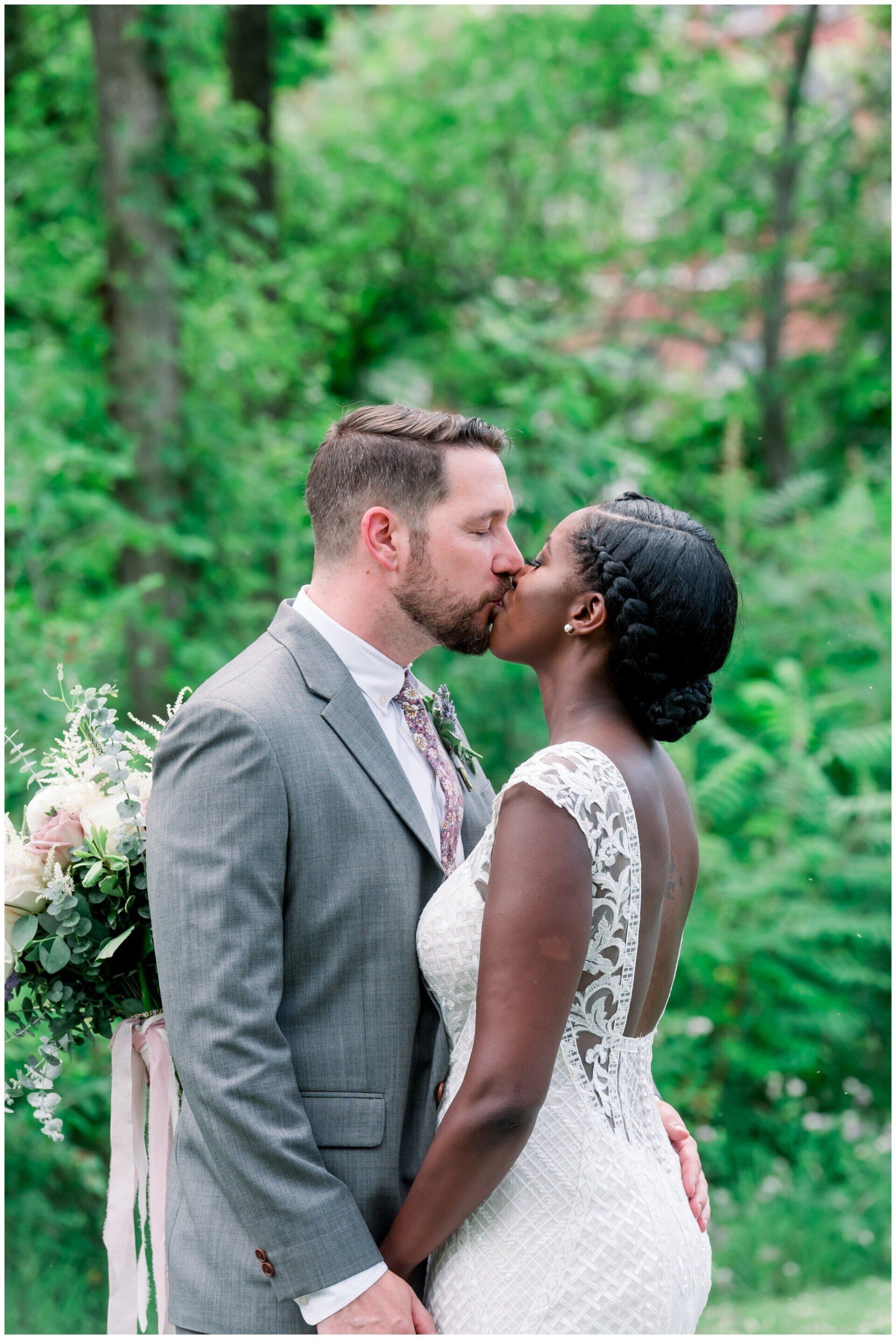 Couple kissing outdoors in a green garden, groom in gray suit and bride in white lace dress