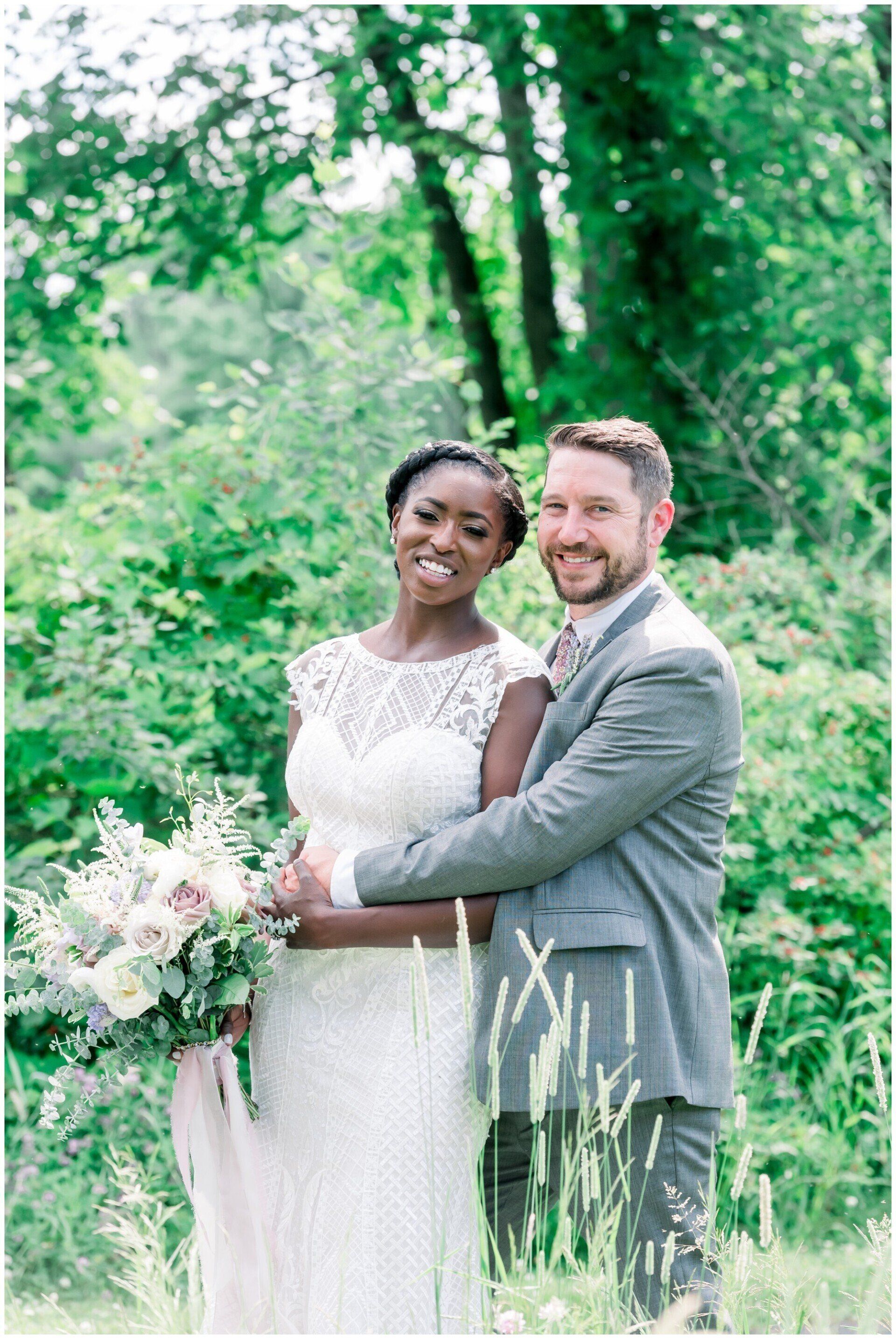 Two people in wedding attire smiling in a green garden, holding a bouquet.