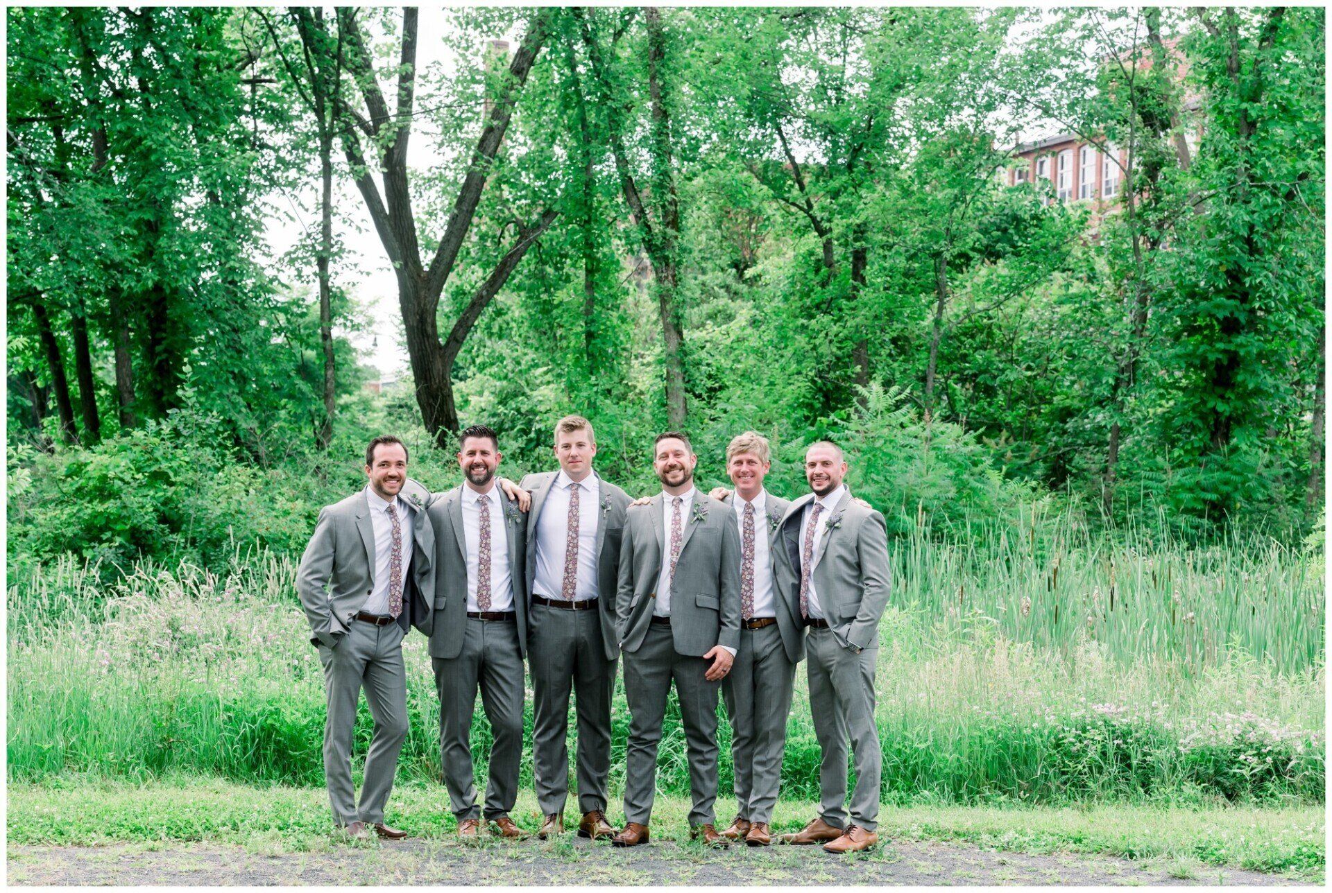Six men in suits standing together on a dirt path in a green wooded area.