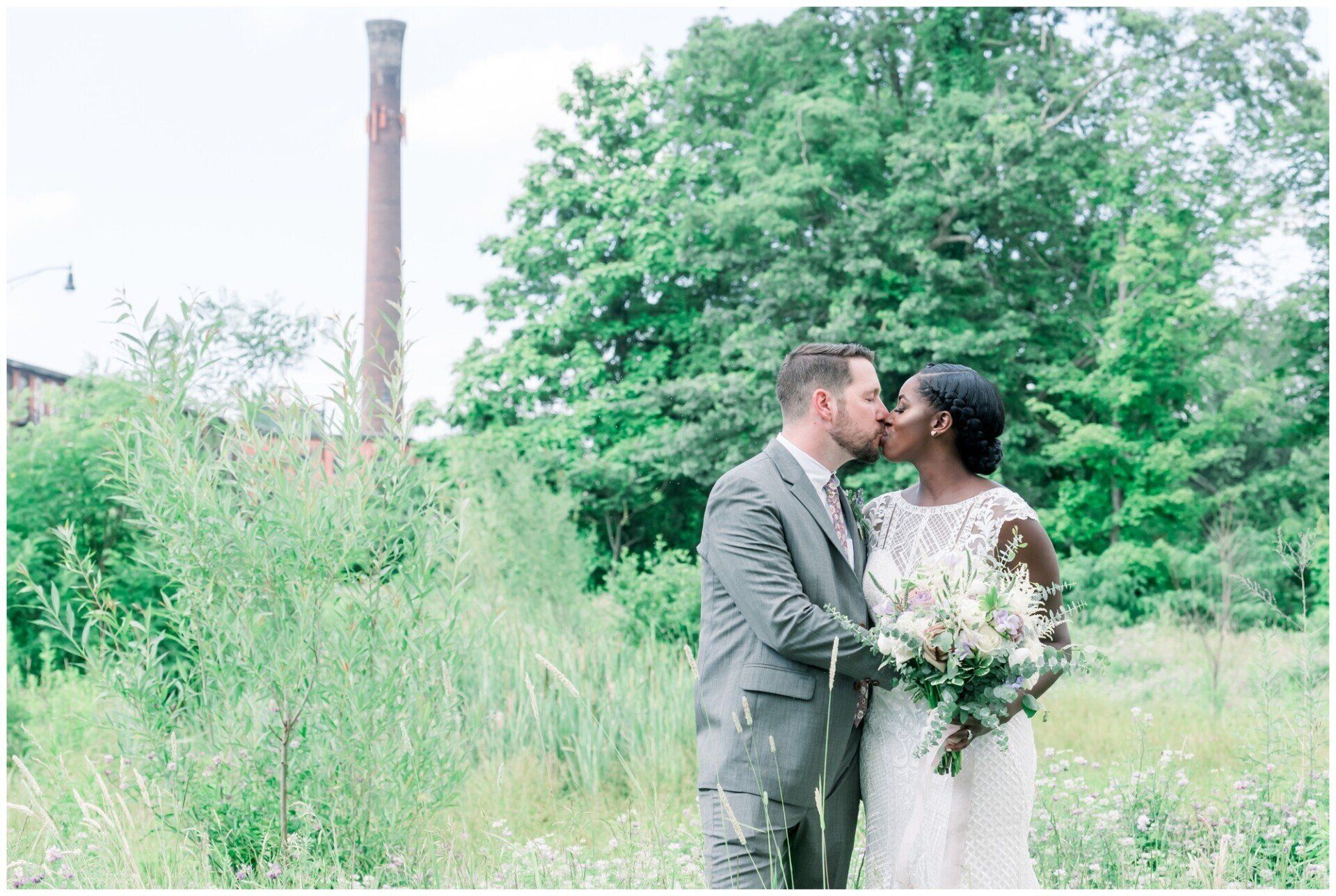 Newlyweds kissing in a grassy field beside tall trees and a brick chimney