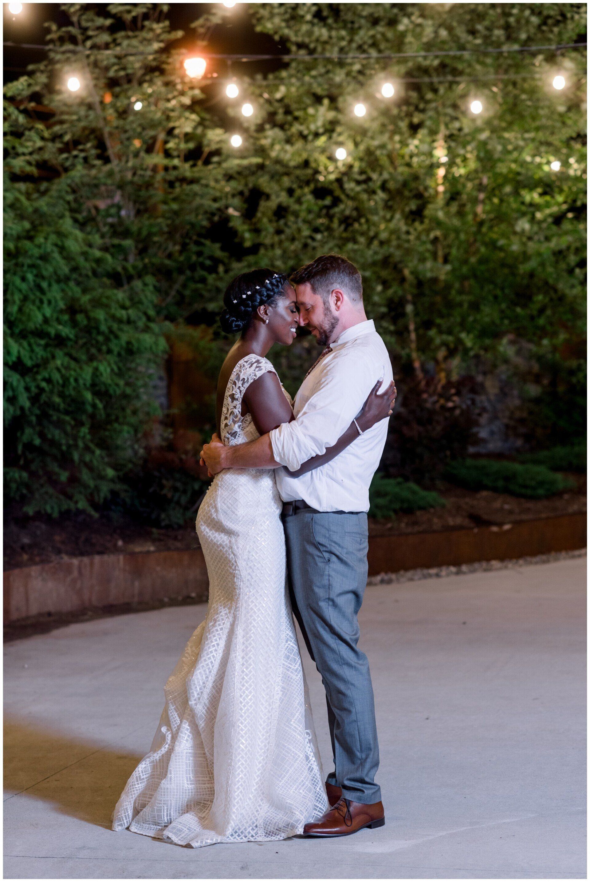 Couple in wedding attire kissing outdoors under string lights at dusk