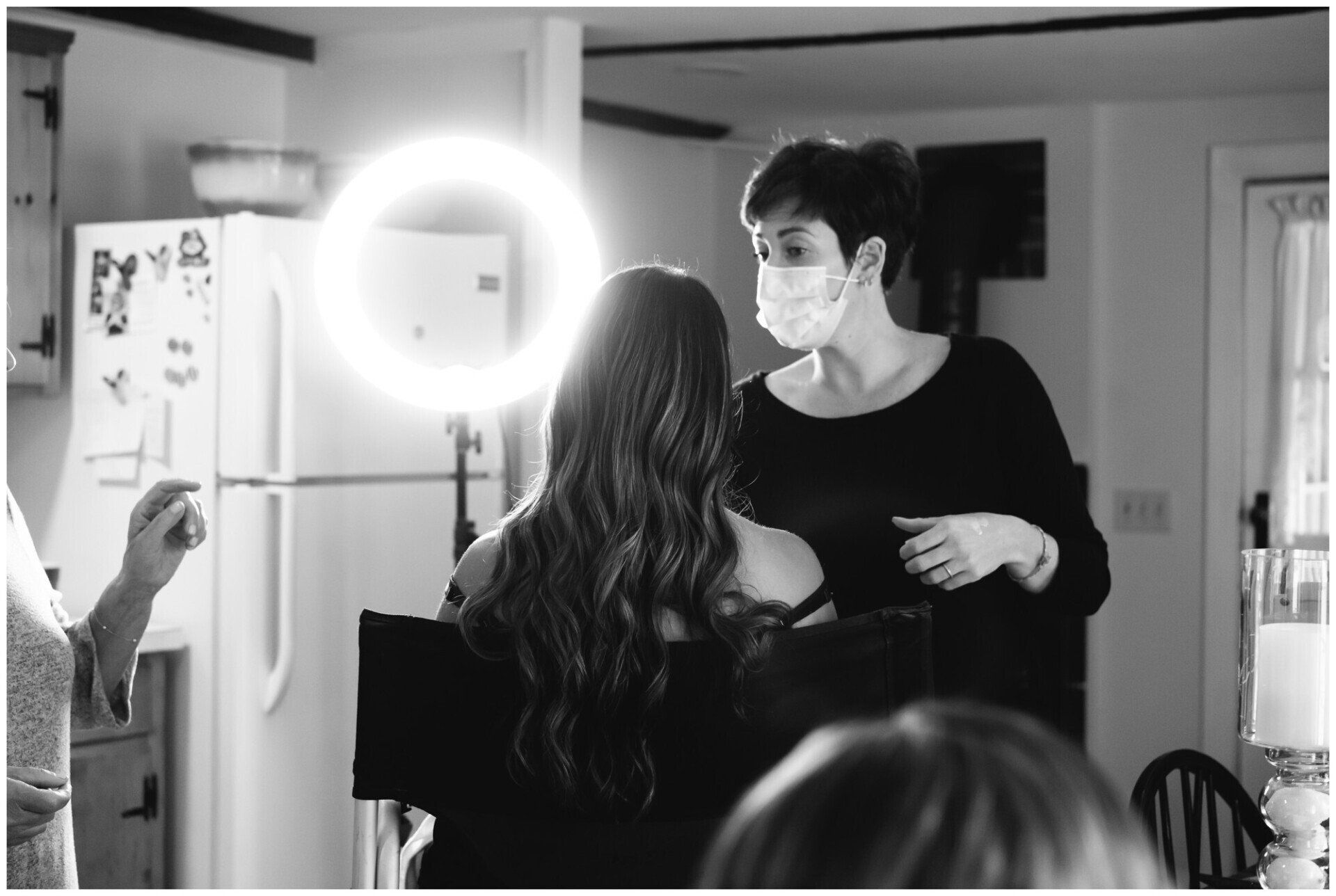Masked stylist gives a haircut in a salon, with a bright ring light nearby.
