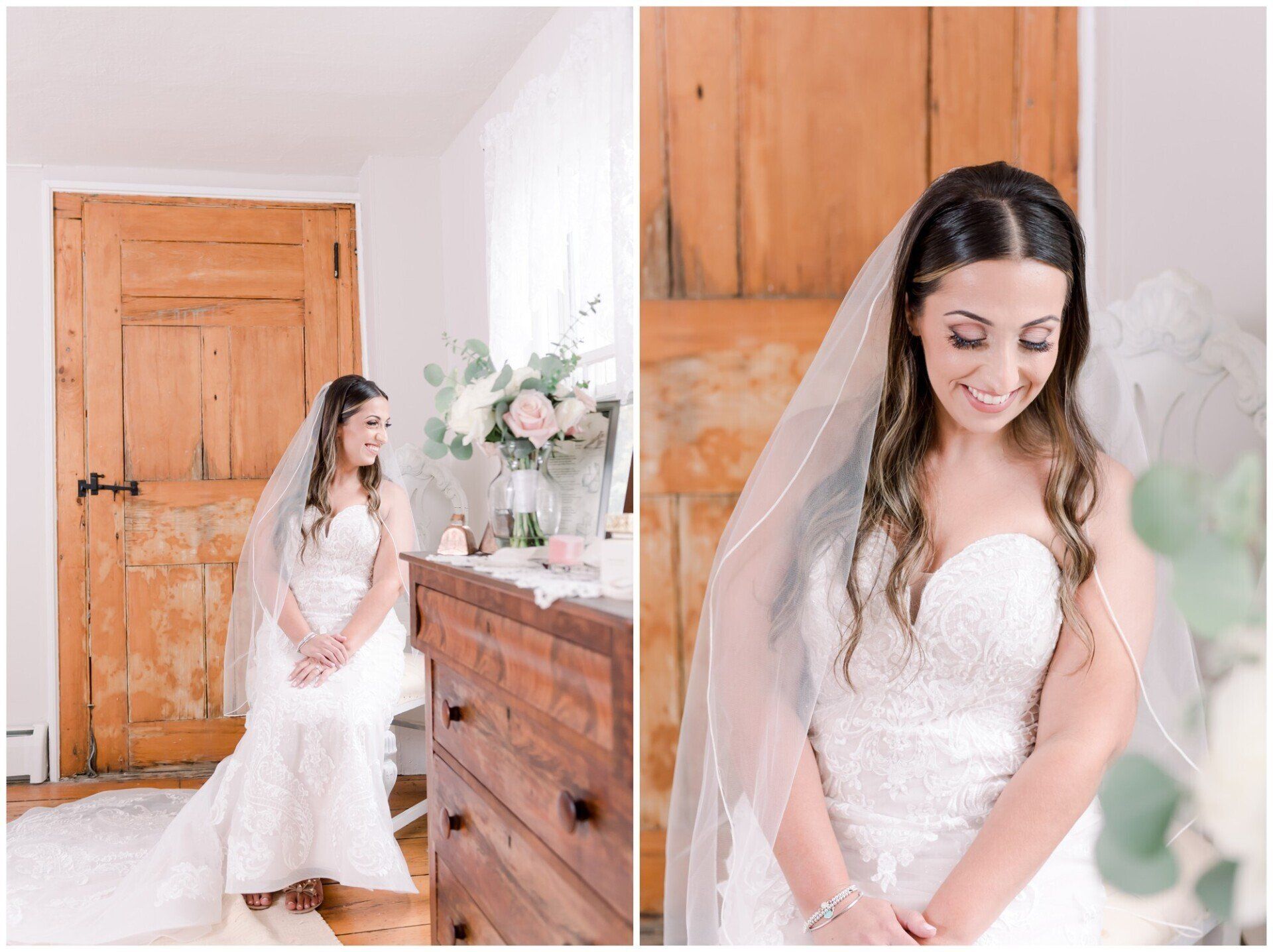 Bride in a white lace dress and veil sitting in a bright rustic room beside a wooden dresser with flowers.