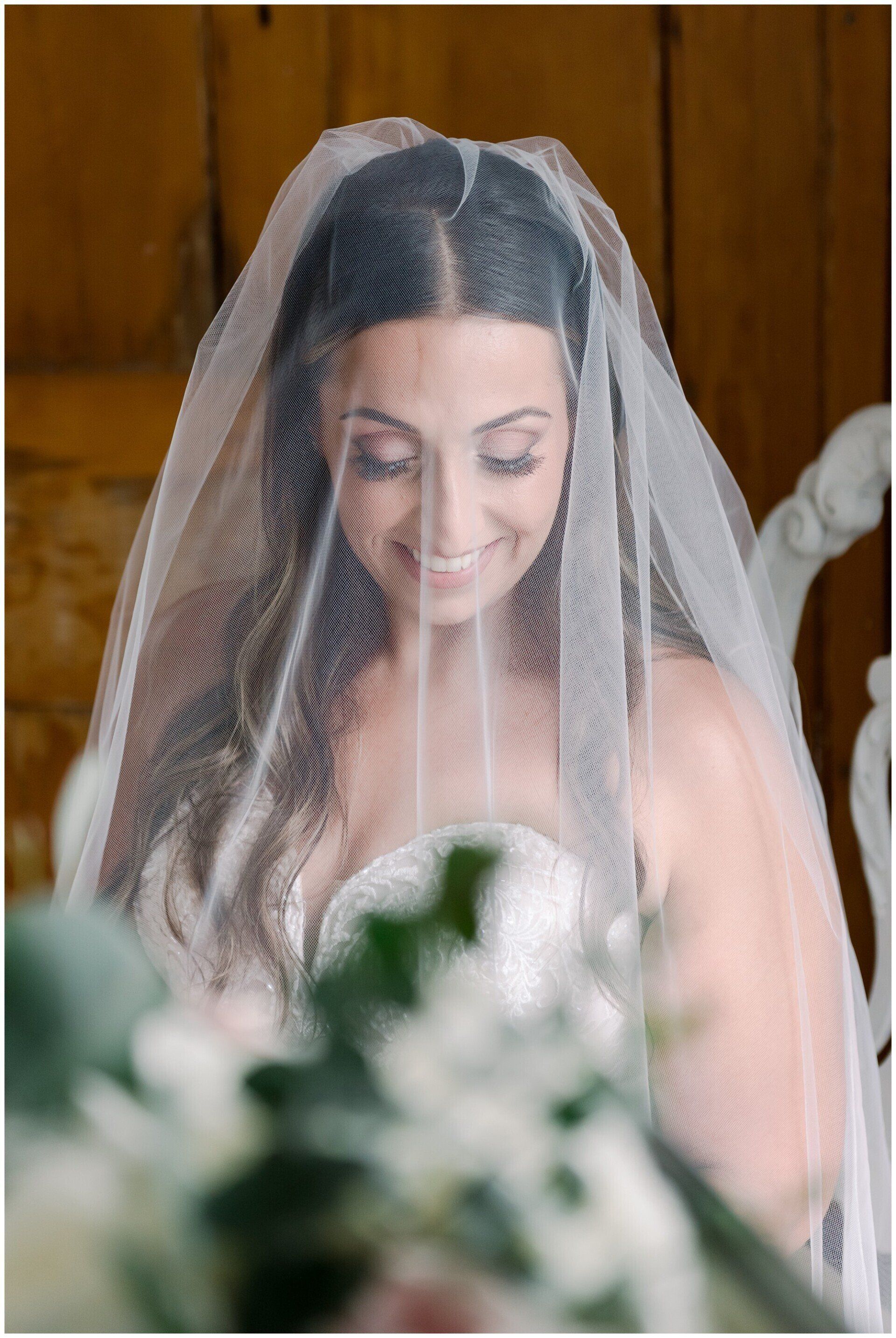 Bride smiling under a white veil, with blurred flowers in the foreground.