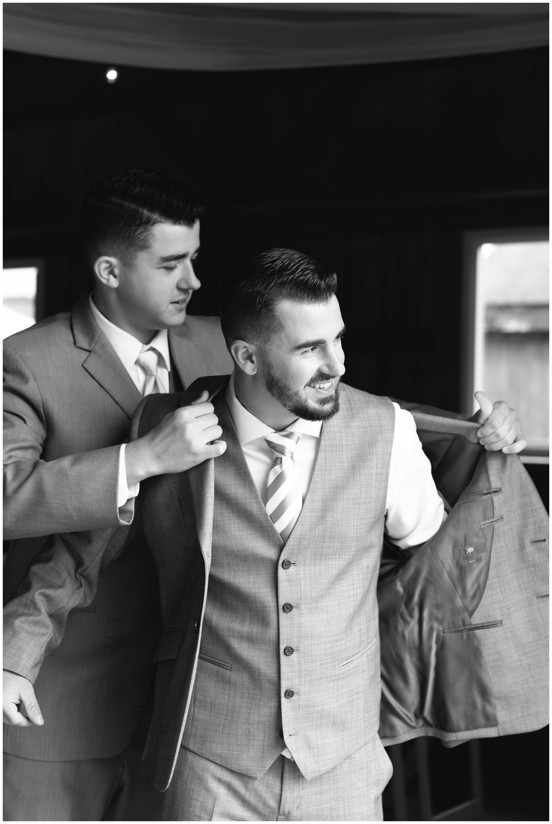 Two men in suits adjusting jackets, one helping the other, in a black-and-white indoor scene