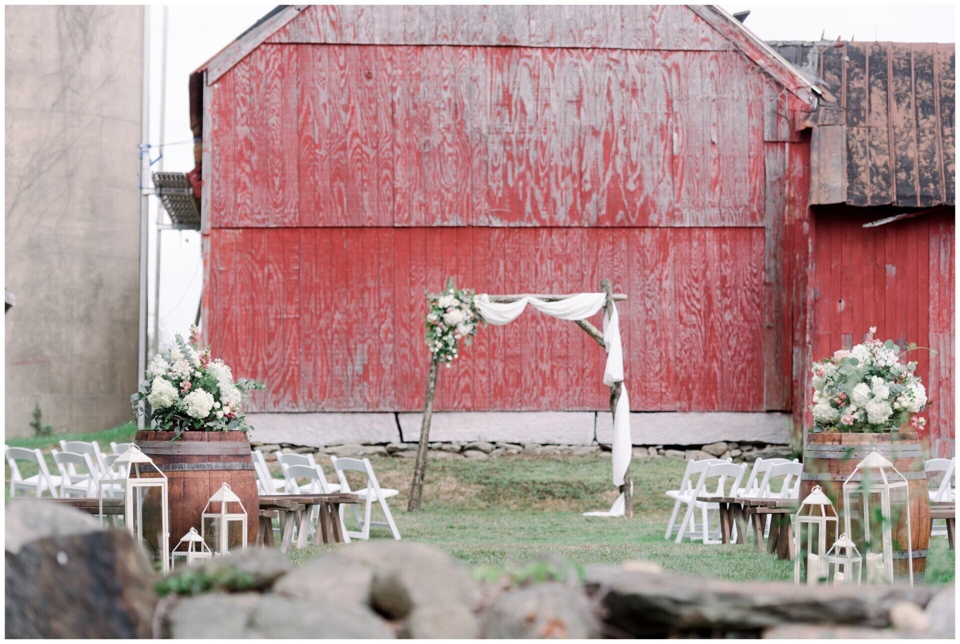 Rustic outdoor wedding setup with white chairs and floral arch before a red barn