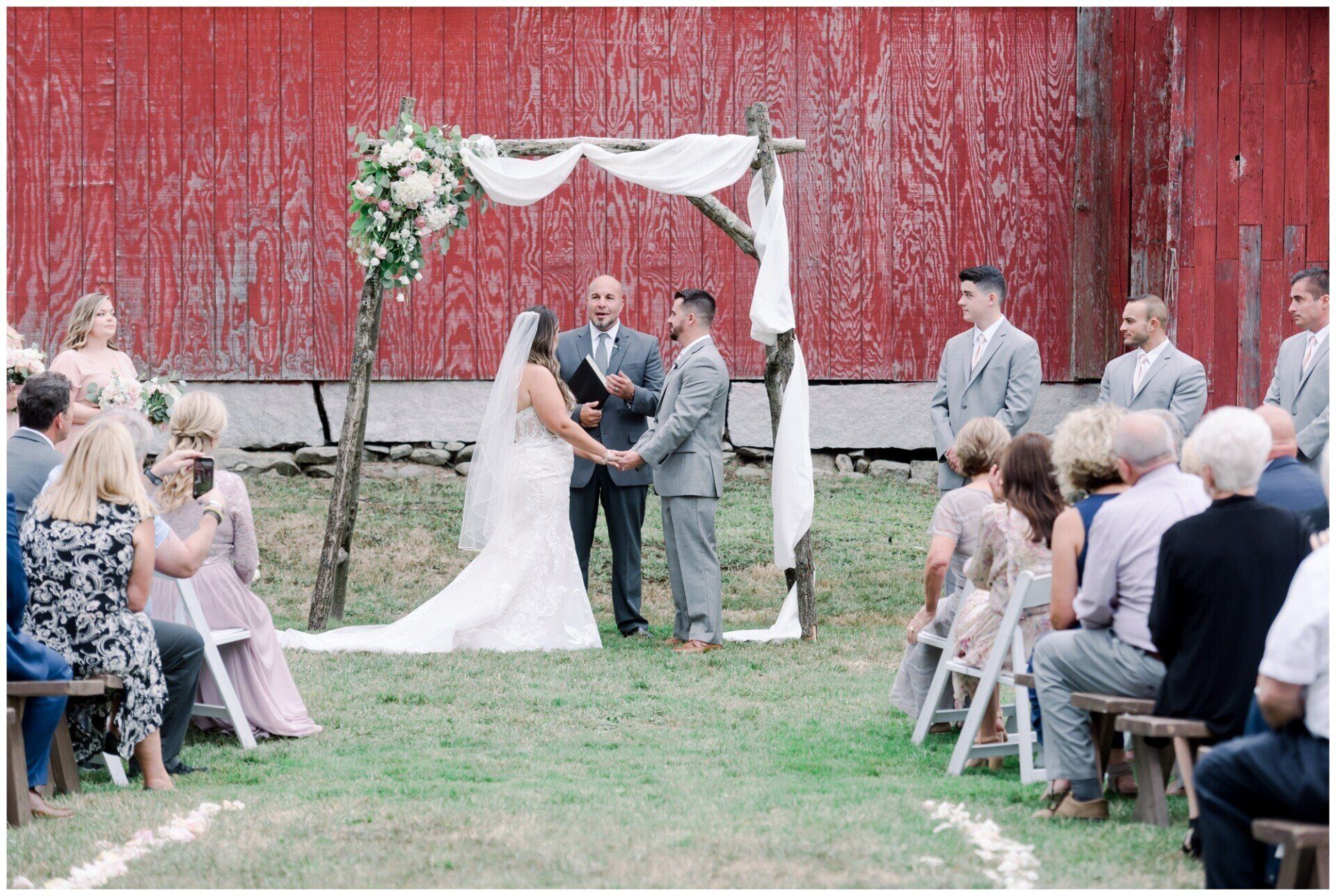Wedding ceremony under a white arch in front of a red barn, with guests seated on both sides.