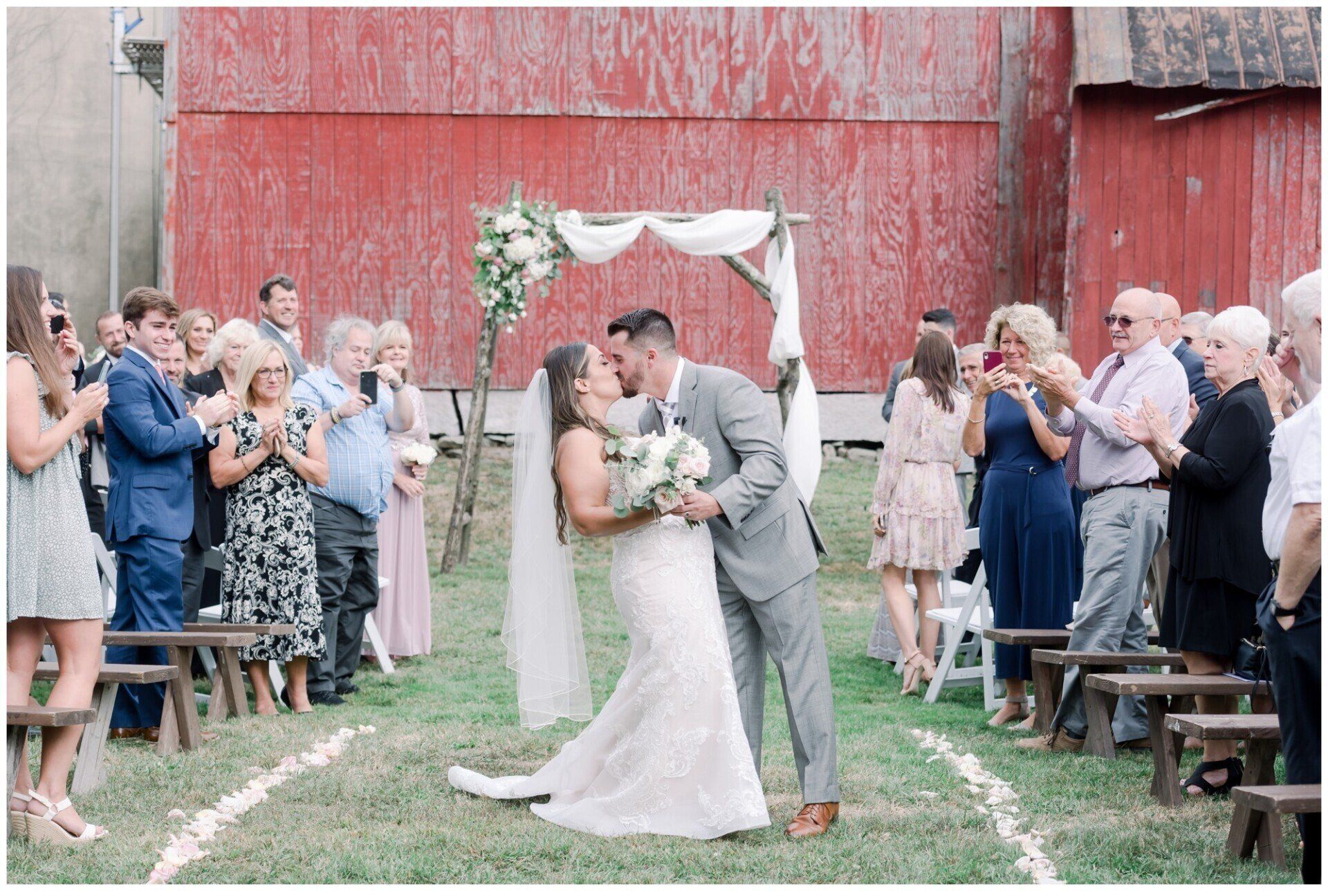 Outdoor wedding kiss beneath a white arch, with guests applauding beside a red barn.
