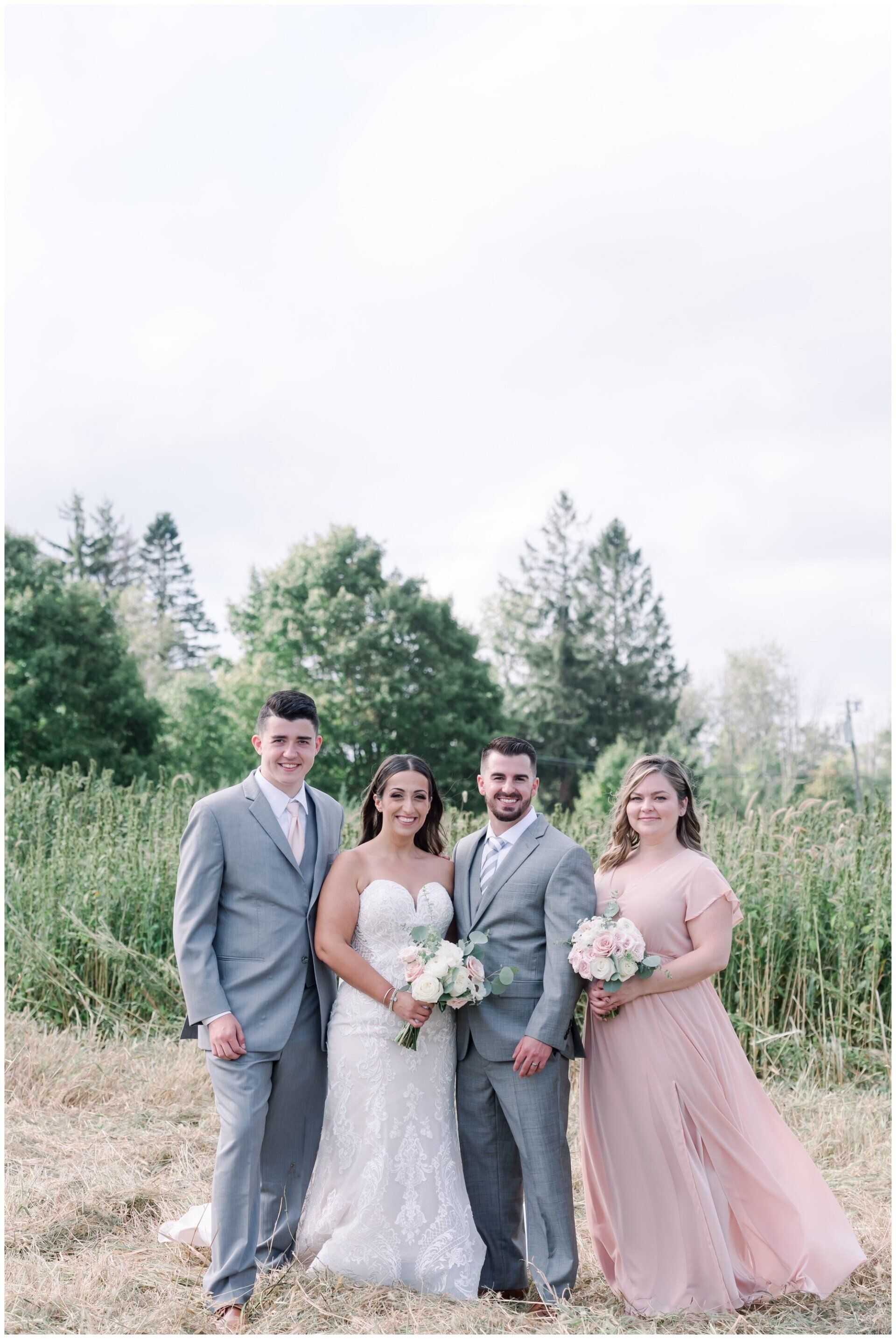 Four wedding party members pose outdoors in formal gray and blush attire, holding bouquets.