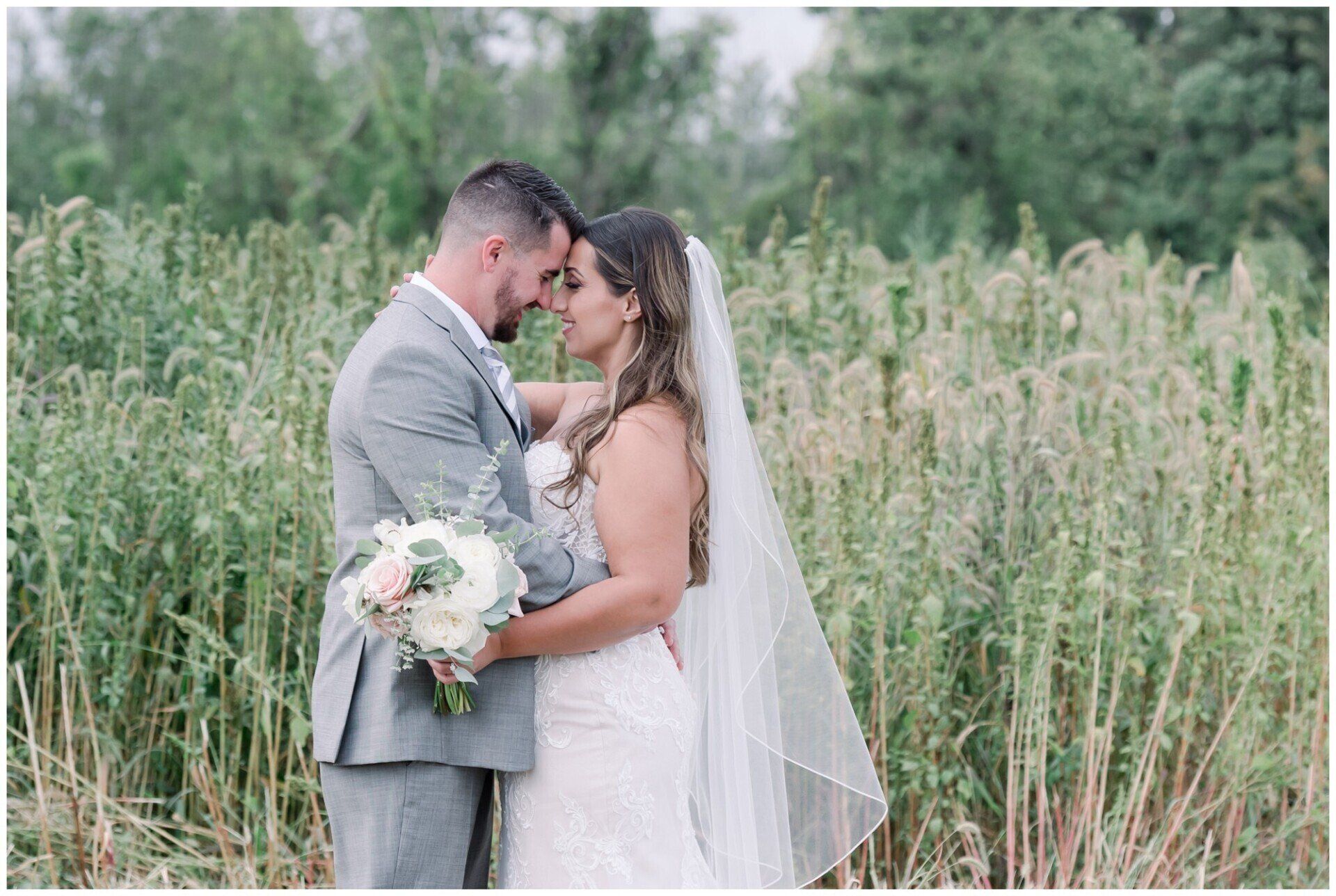 Bride and groom embracing in a grassy field, holding a bouquet and wearing wedding attire.
