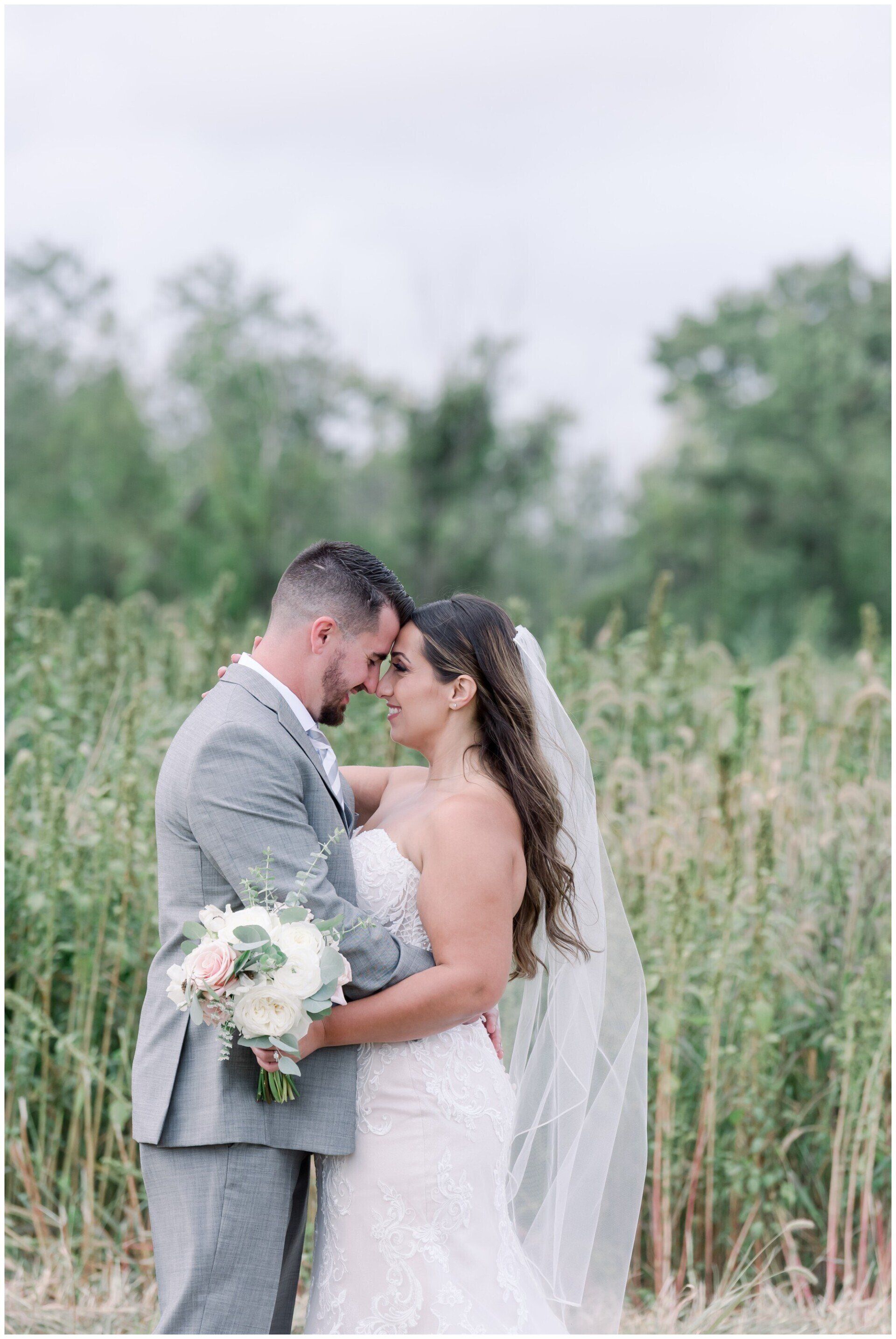 Bride and groom embracing in a grassy field, holding a bouquet, with trees in the background