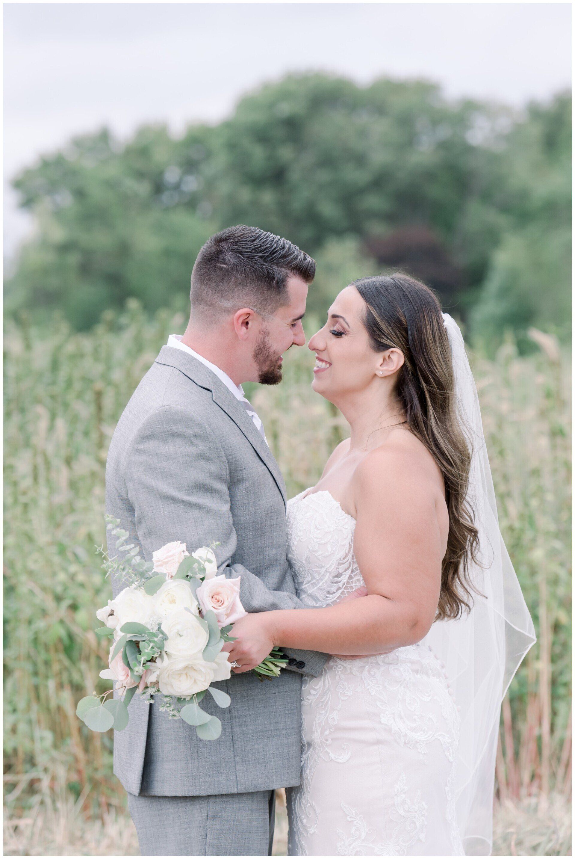 Bride and groom holding hands and facing each other in a grassy field, with bouquets and greenery behind them