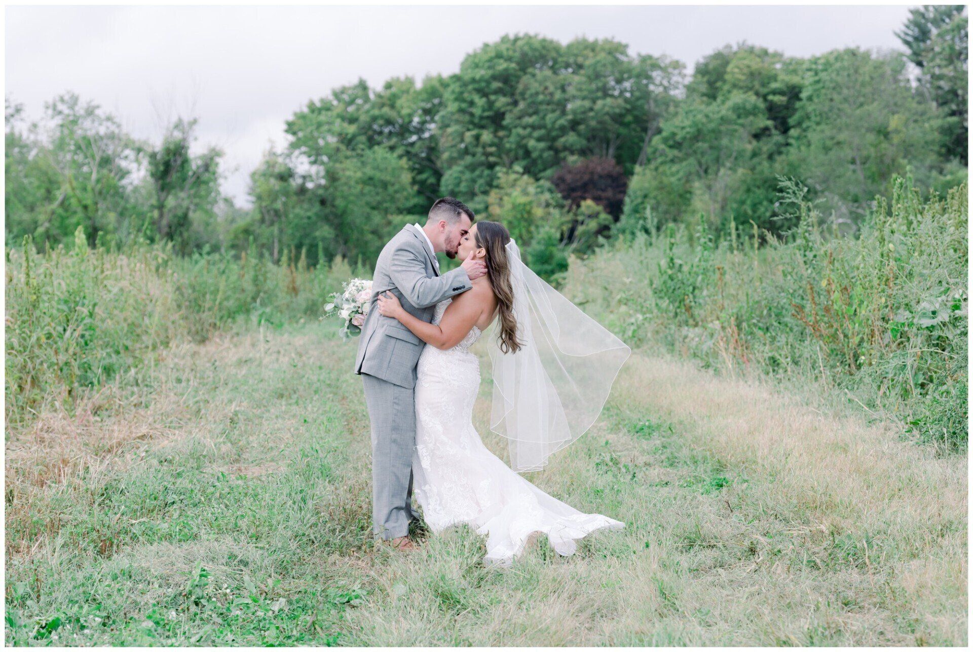 Couple embracing in a grassy field, bride in white dress and veil, groom in gray suit, with trees behind them