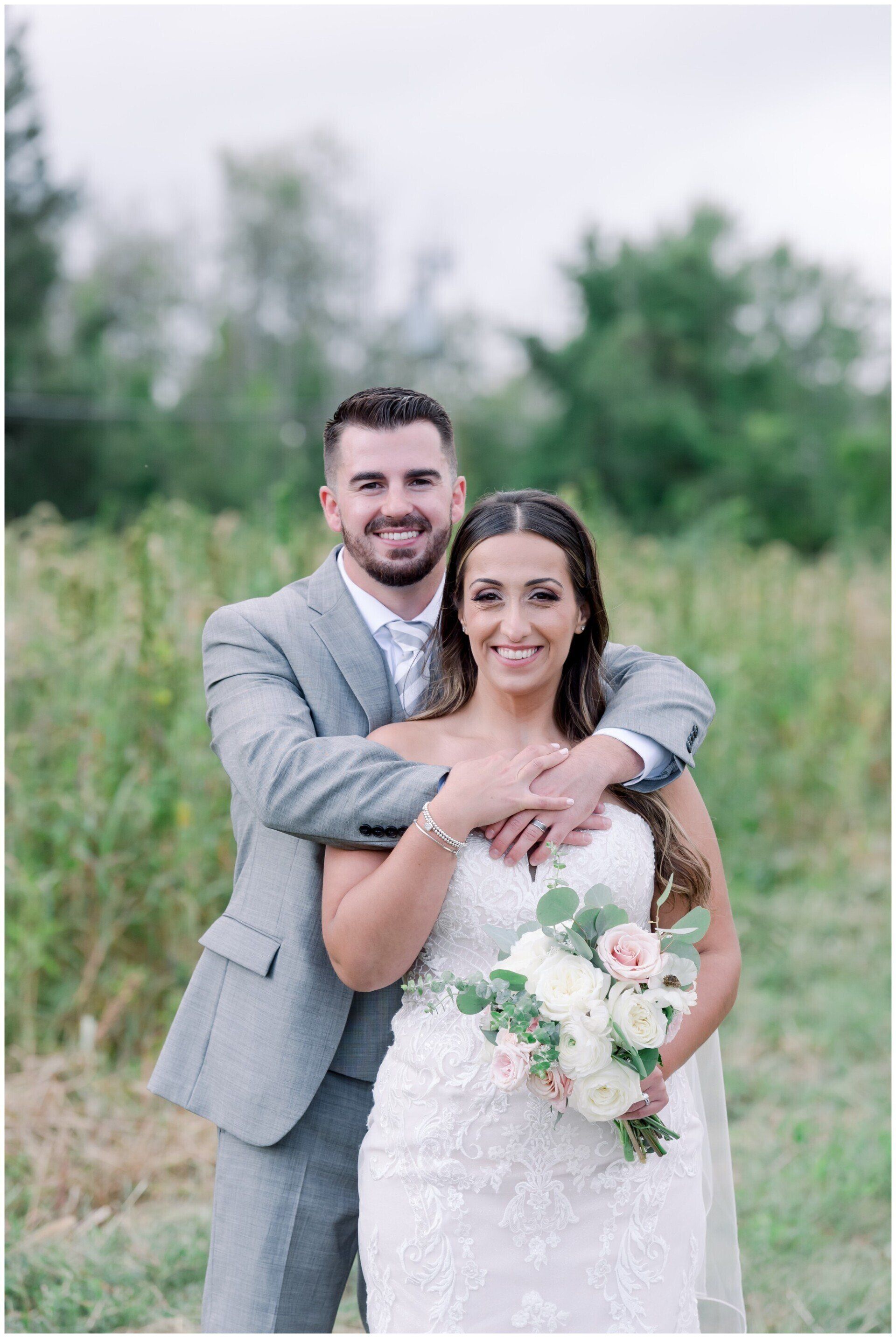 Bride and groom posing outdoors, holding a bouquet, with green trees in the background