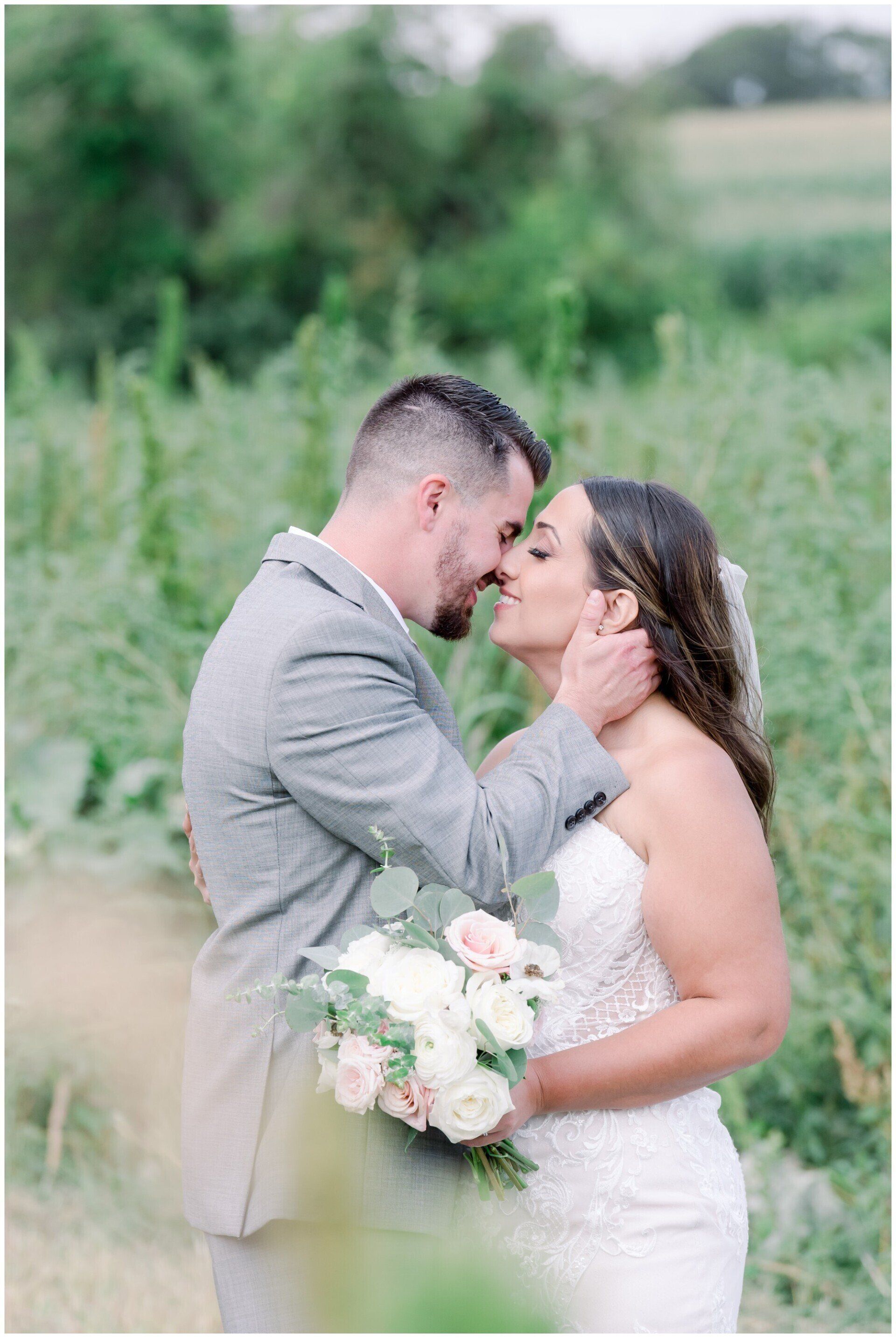 Couple embracing outdoors in a green field, bride holding a white bouquet