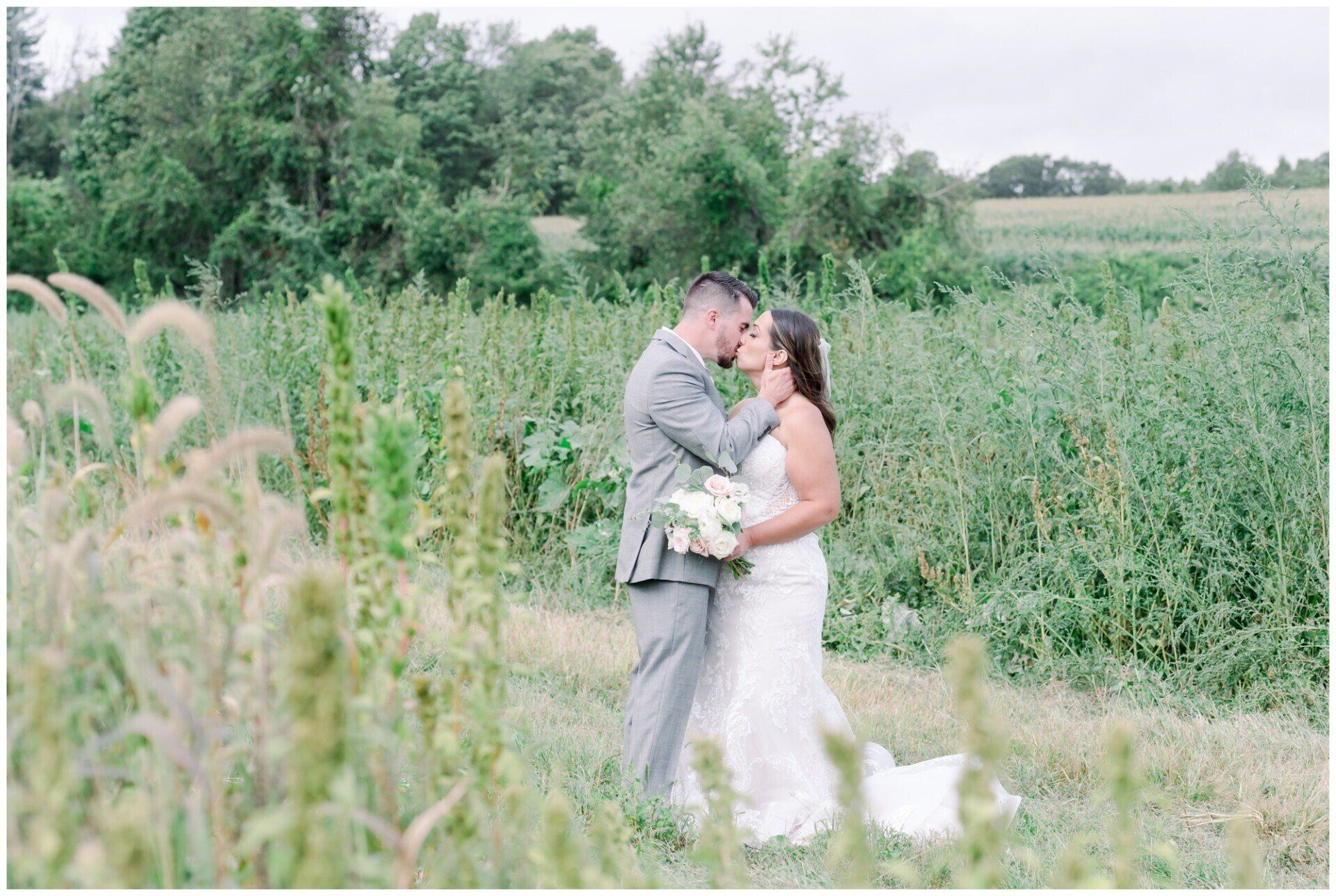 Bride and groom kissing in a grassy field with wildflowers and trees in the background