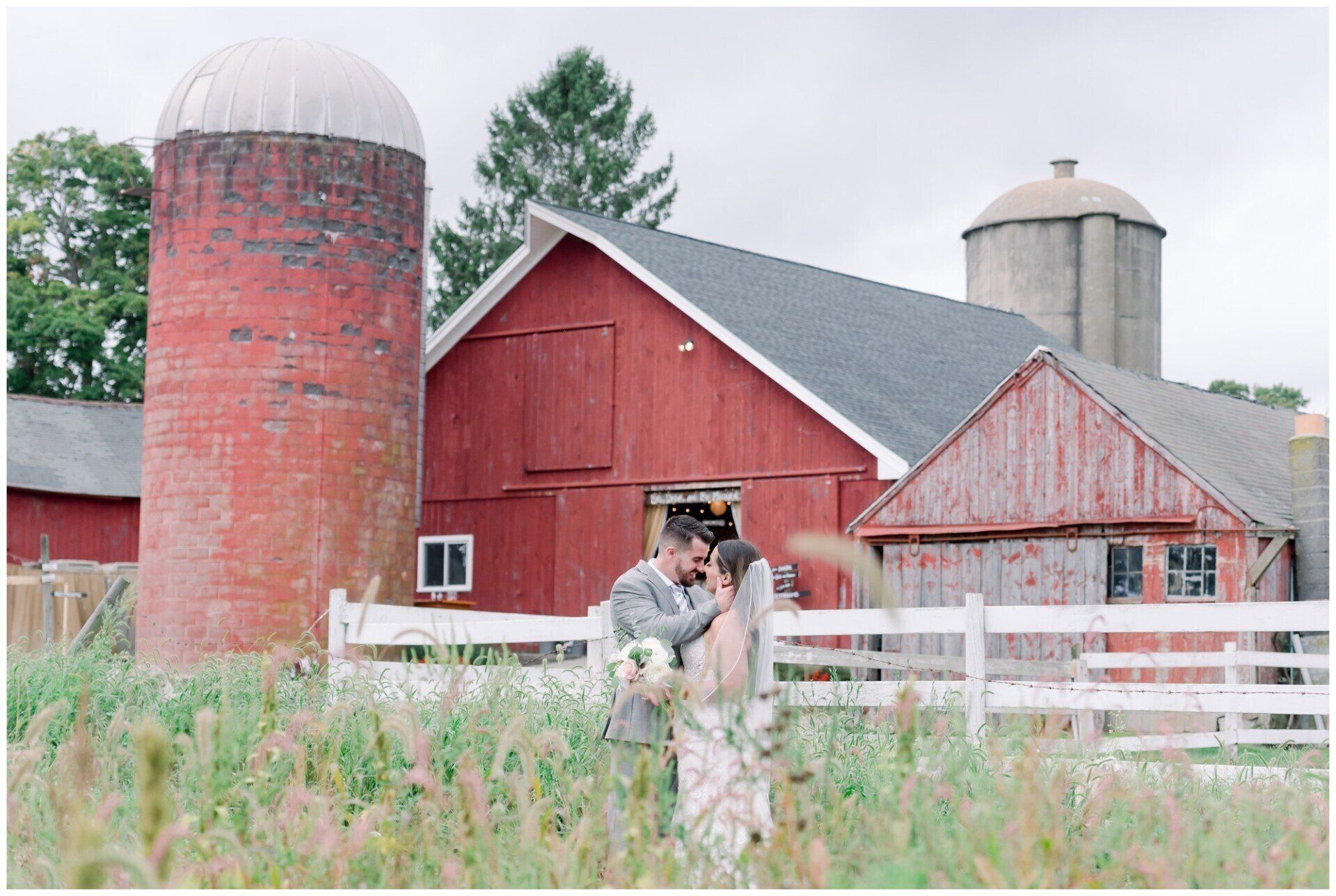 People in a white fence by red barns and silos on a farm field