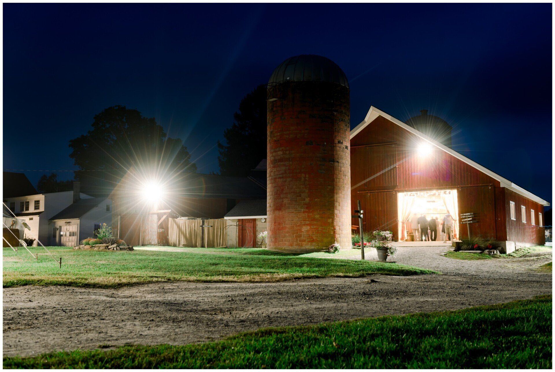 Nighttime farmyard with a red barn, silo, bright lights, and green grass.