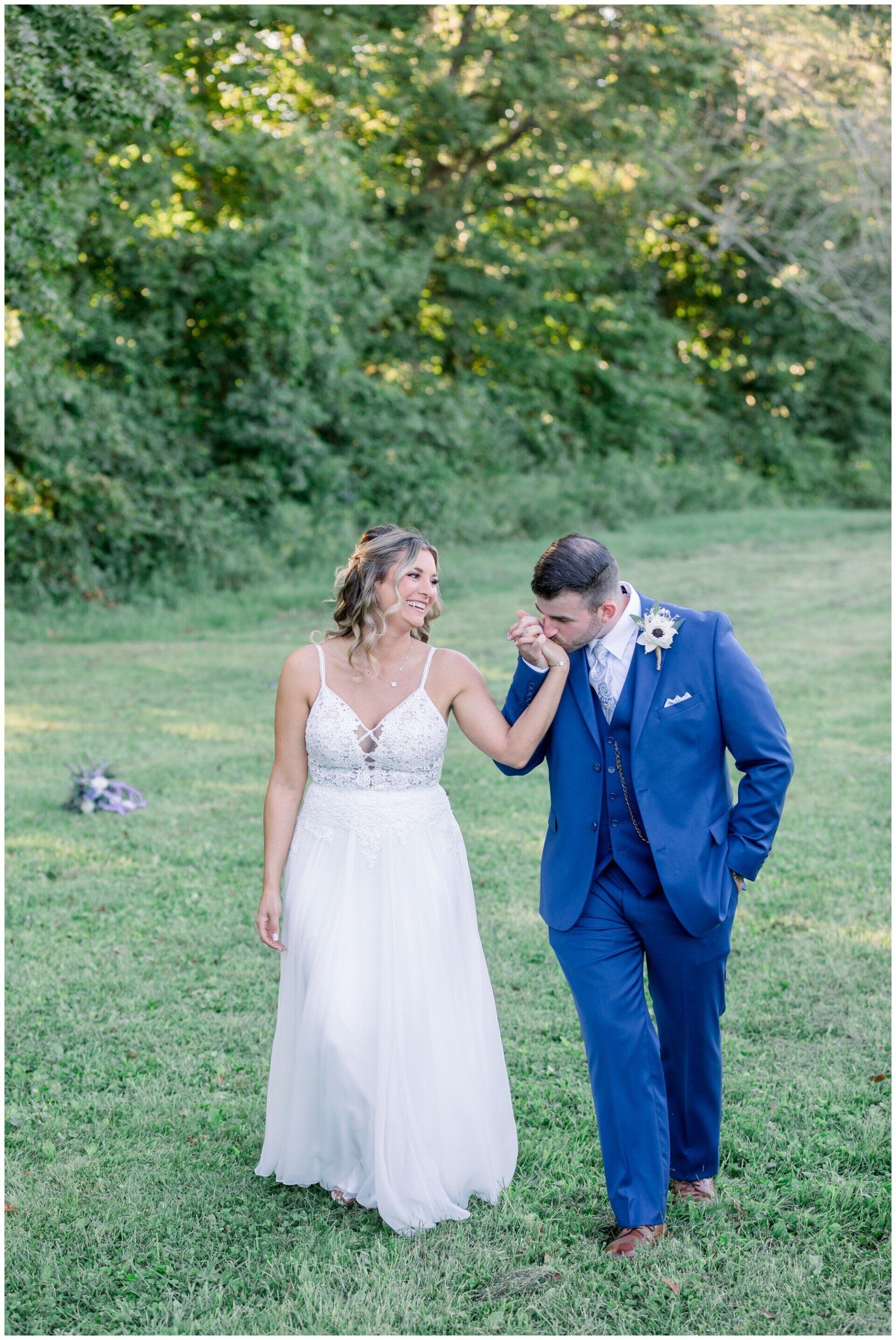Bride and groom holding hands in a grassy field, smiling at each other; the groom wears a blue suit.