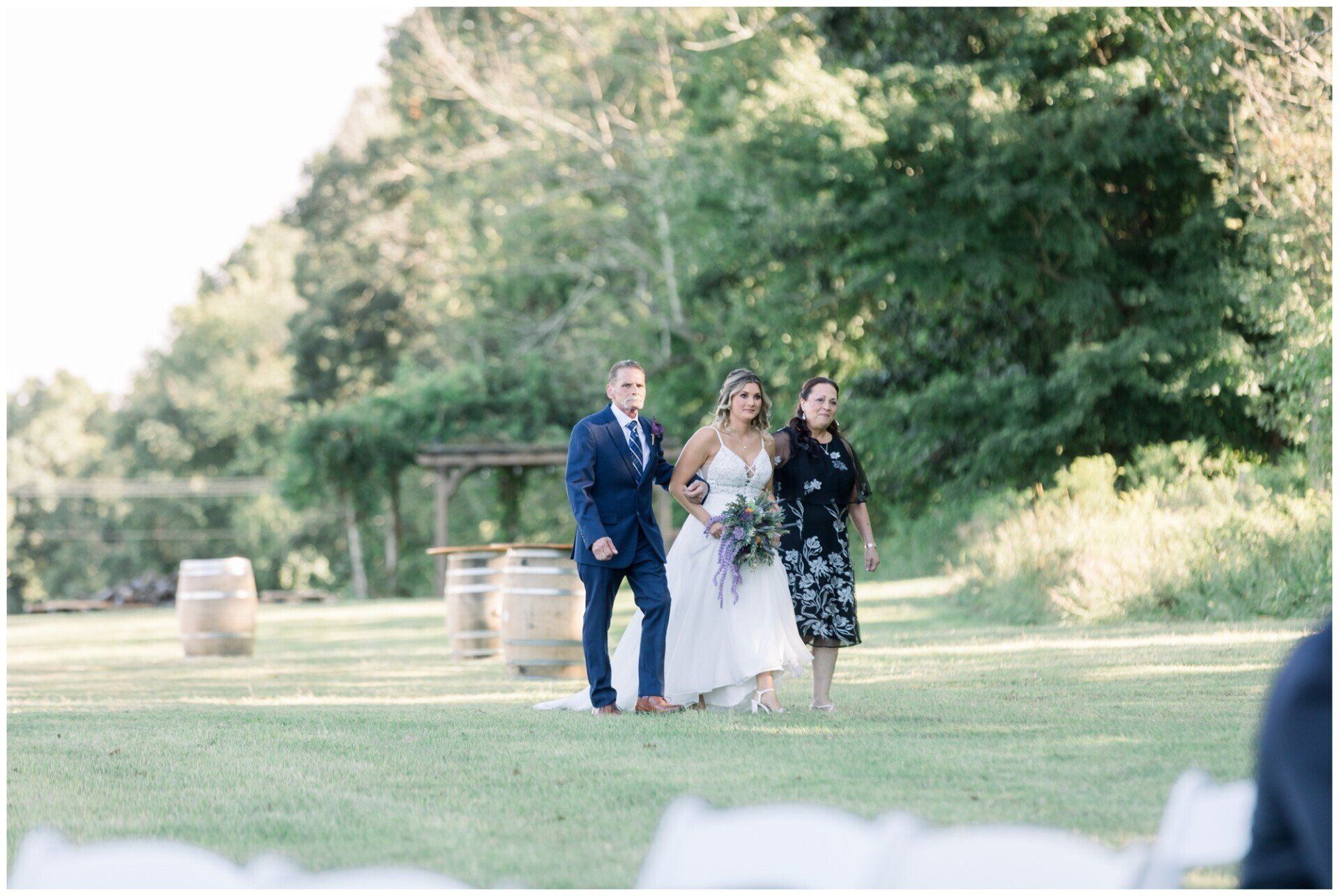 Three people walk across a grassy field at an outdoor wedding, with one in a white dress and the others in dark attire.