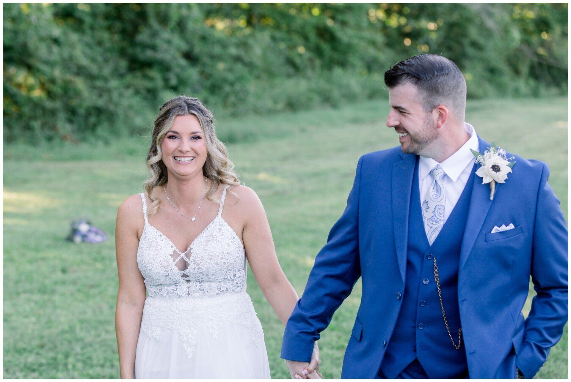 Bride and groom walking hand in hand outdoors, smiling in a grassy field.