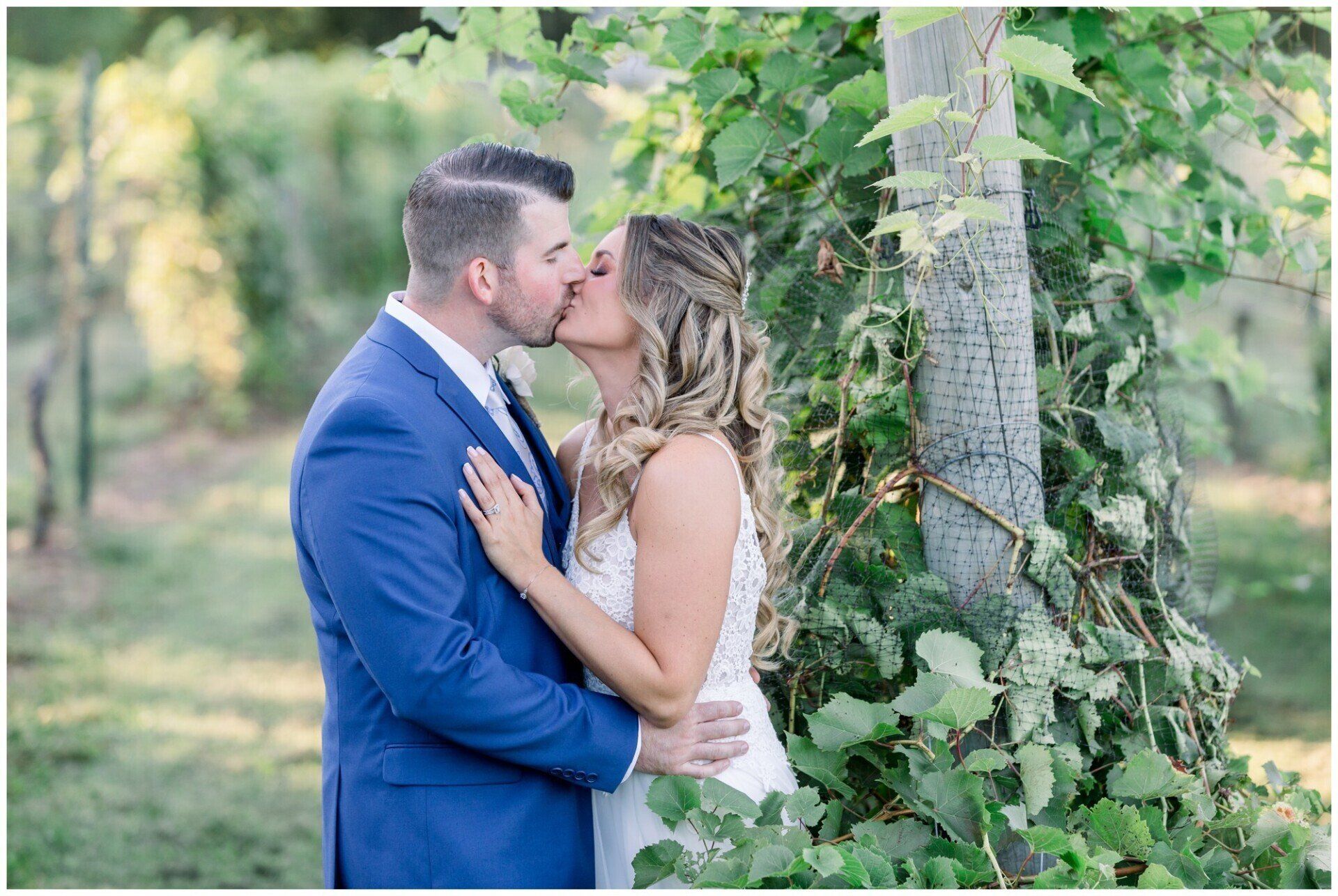 Couple kissing beside a tree in a leafy outdoor setting, the man in a blue suit and the woman in a white dress