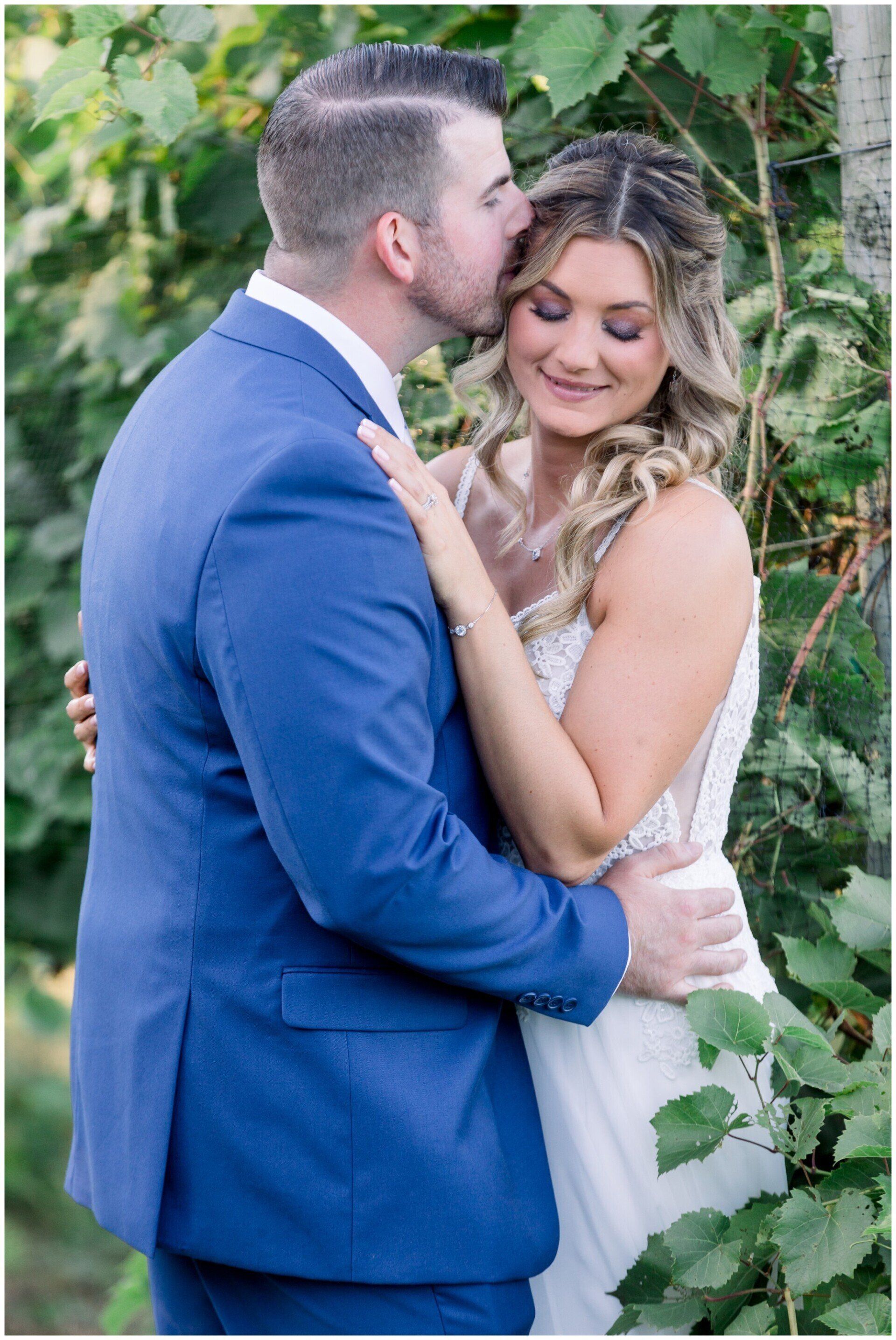 Couple embracing outdoors, man in blue suit kissing woman in white dress amid greenery