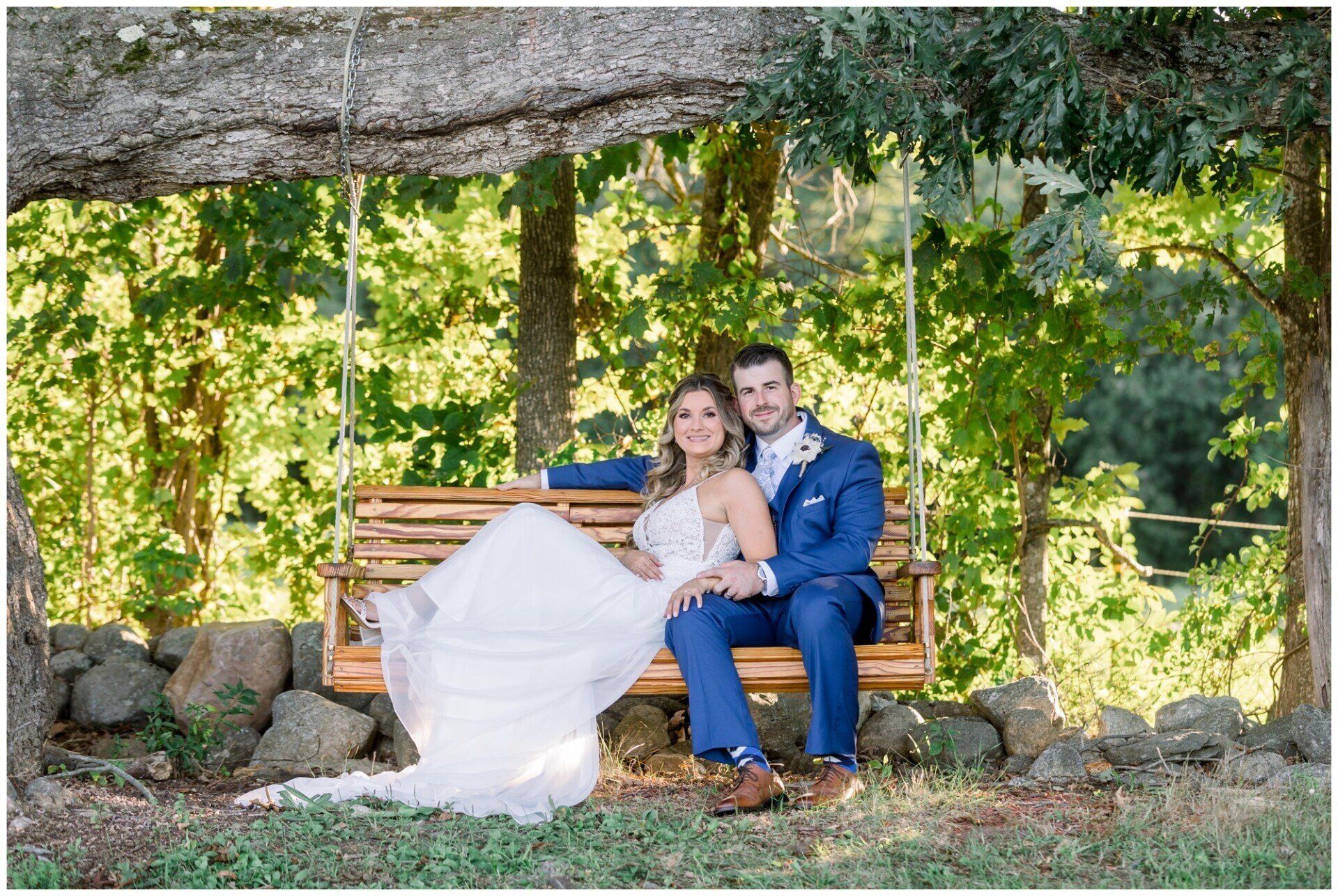 Couple seated on a wooden swing under a tree in a sunny garden, with greenery and stone backdrop