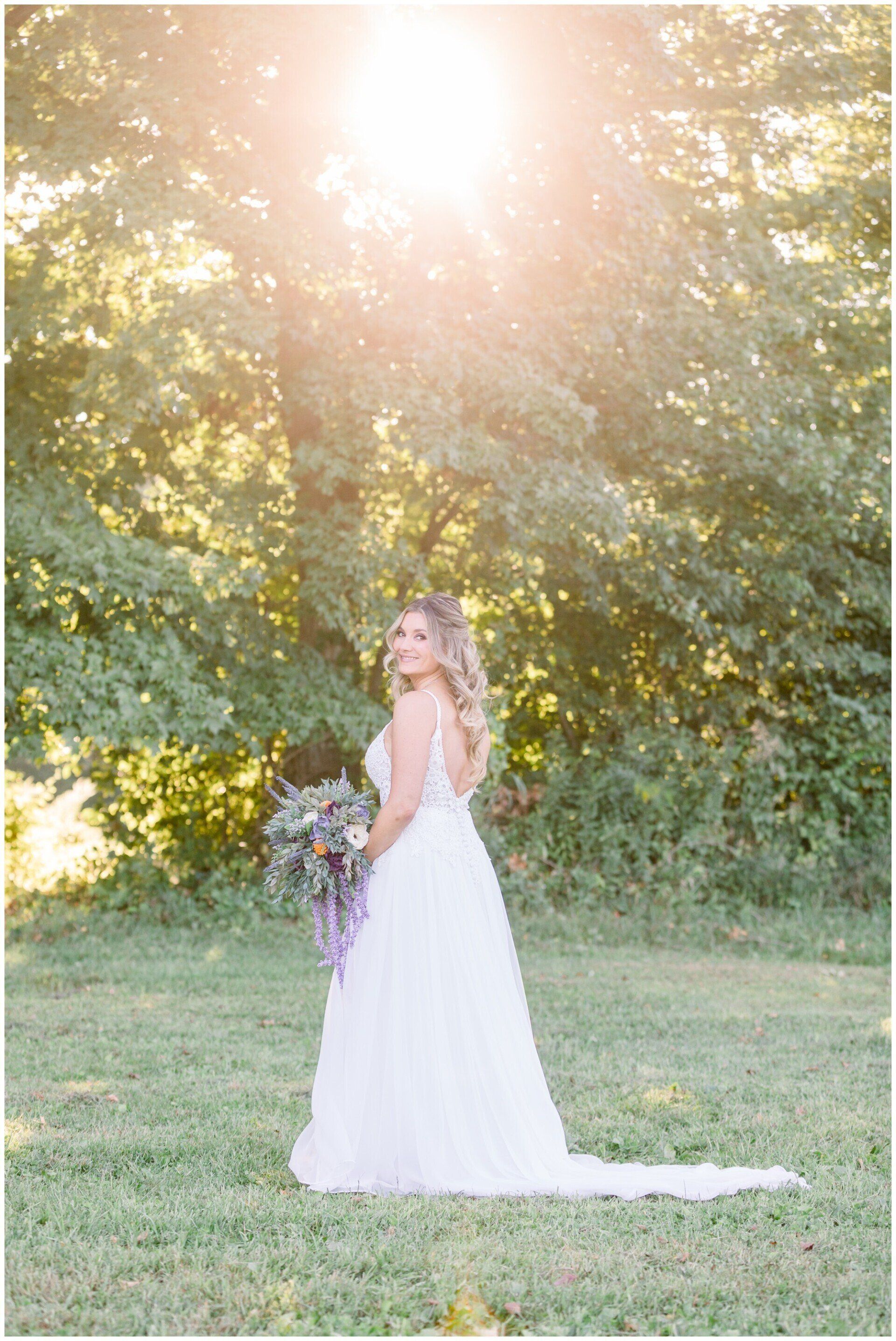 Bride in a white dress holding a bouquet in a sunlit grassy clearing by trees