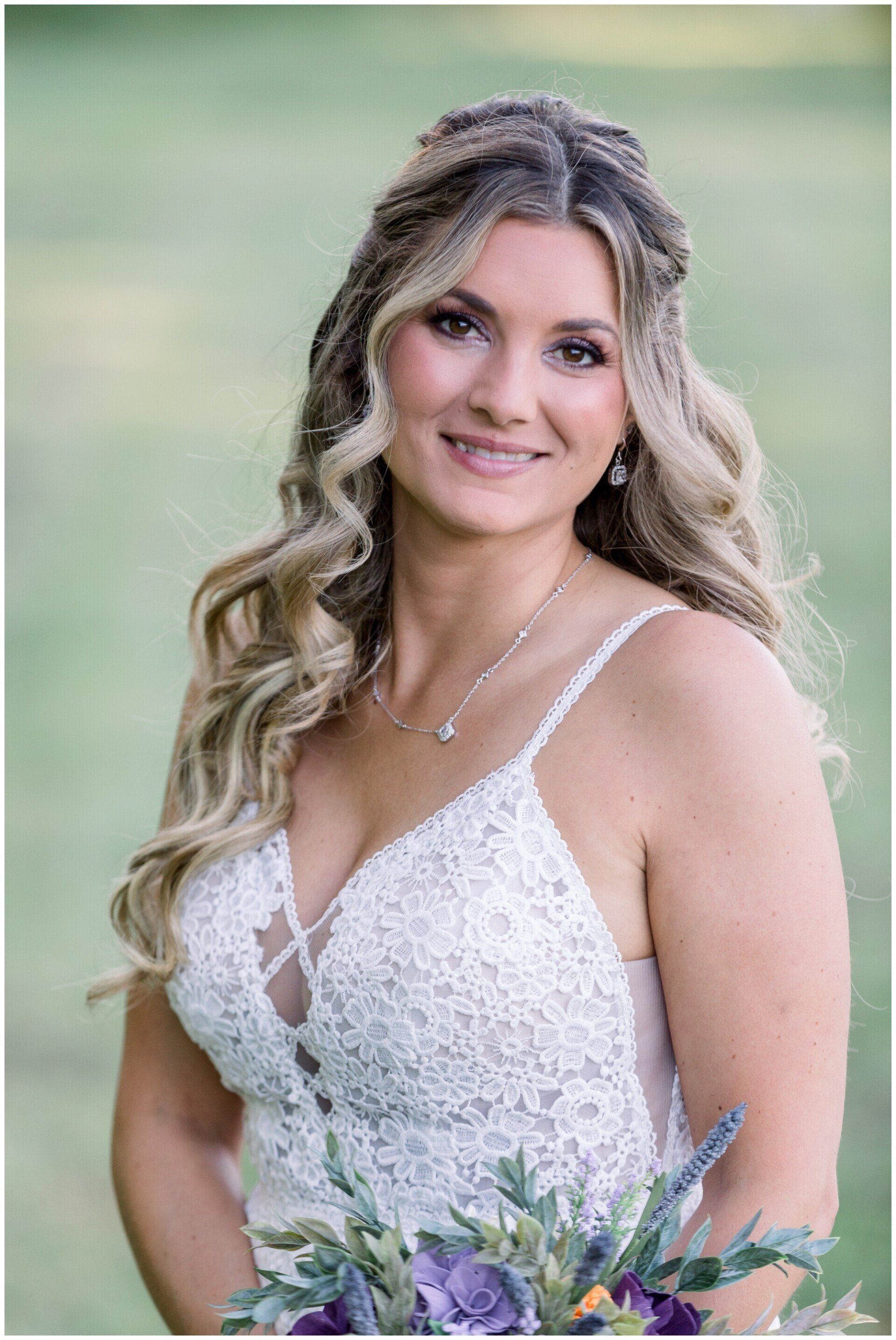Smiling person in a white lace dress holding a purple bouquet outdoors against a soft green background