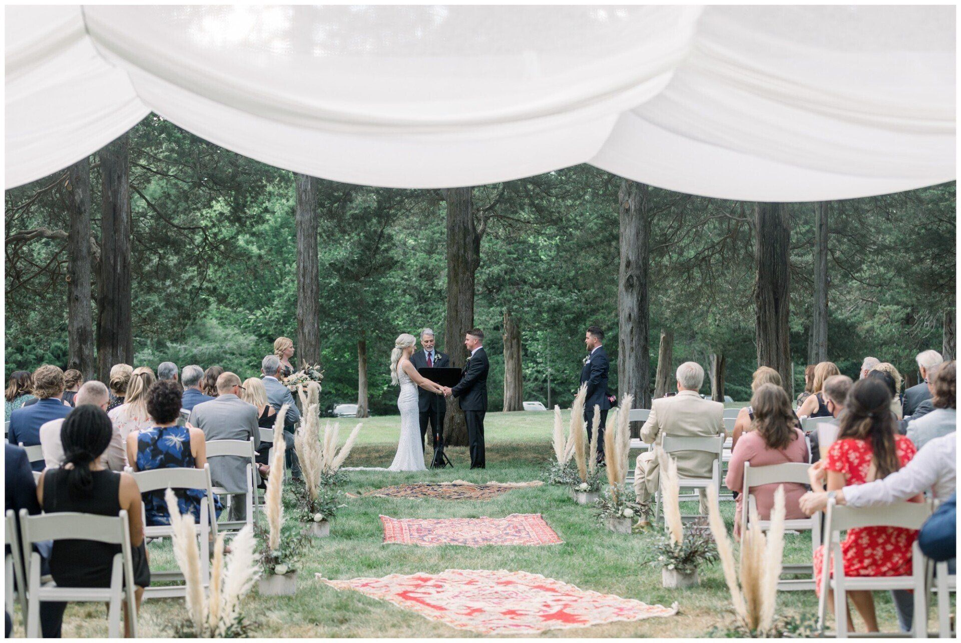 Outdoor wedding ceremony under a white canopy in a wooded clearing, guests seated on both sides of aisle