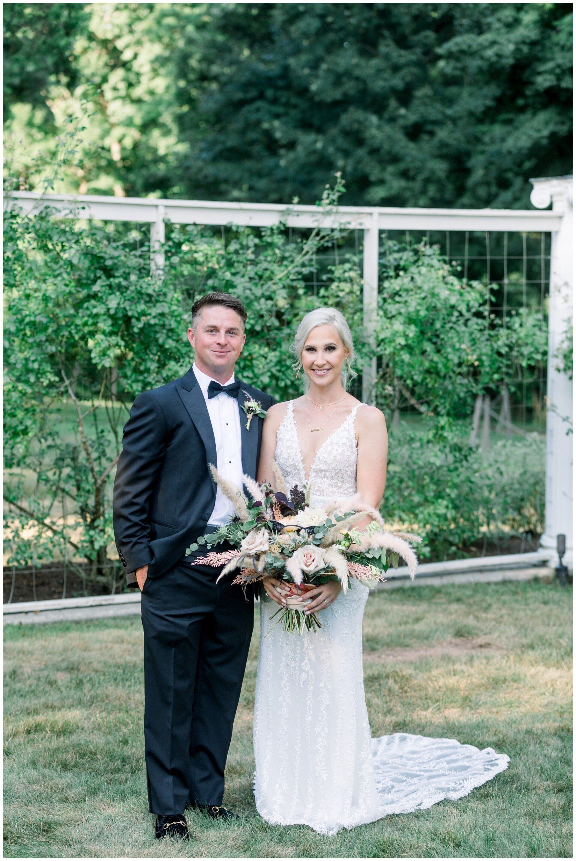 Bride and groom posing outdoors in front of a garden fence, holding a bouquet