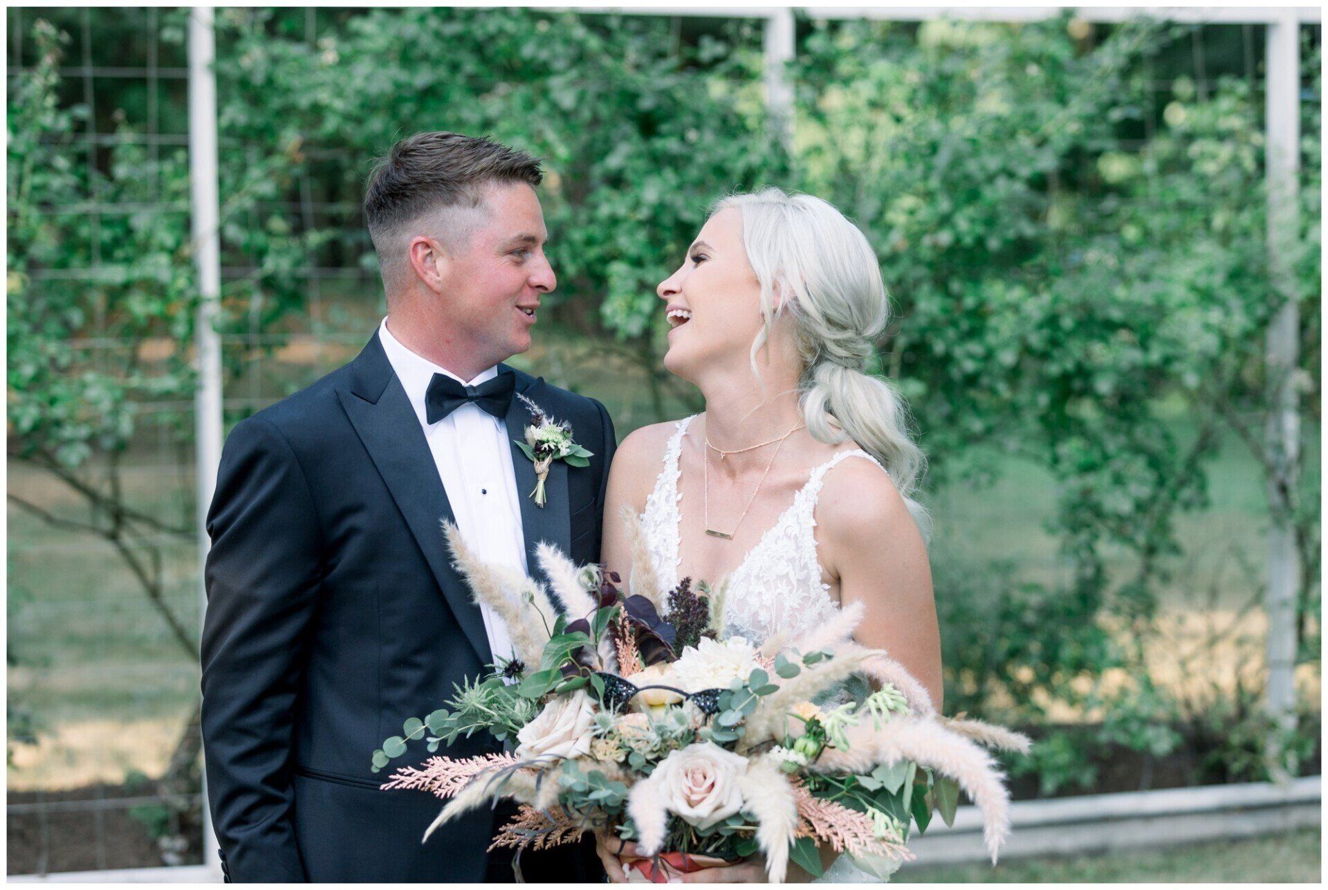 Bride and groom smiling at each other, holding a bouquet, outdoors in a garden setting