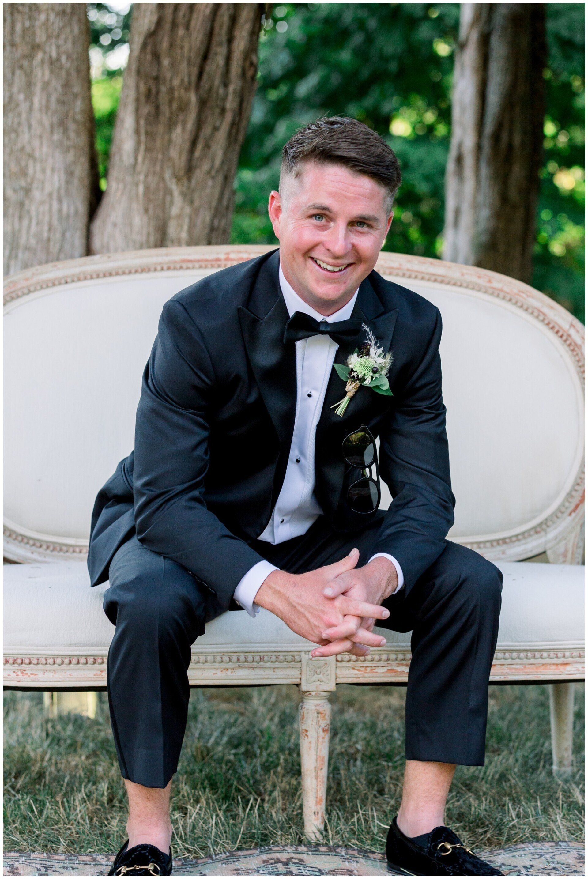 Man in a black suit sitting on a cream sofa outdoors, smiling with hands clasped.