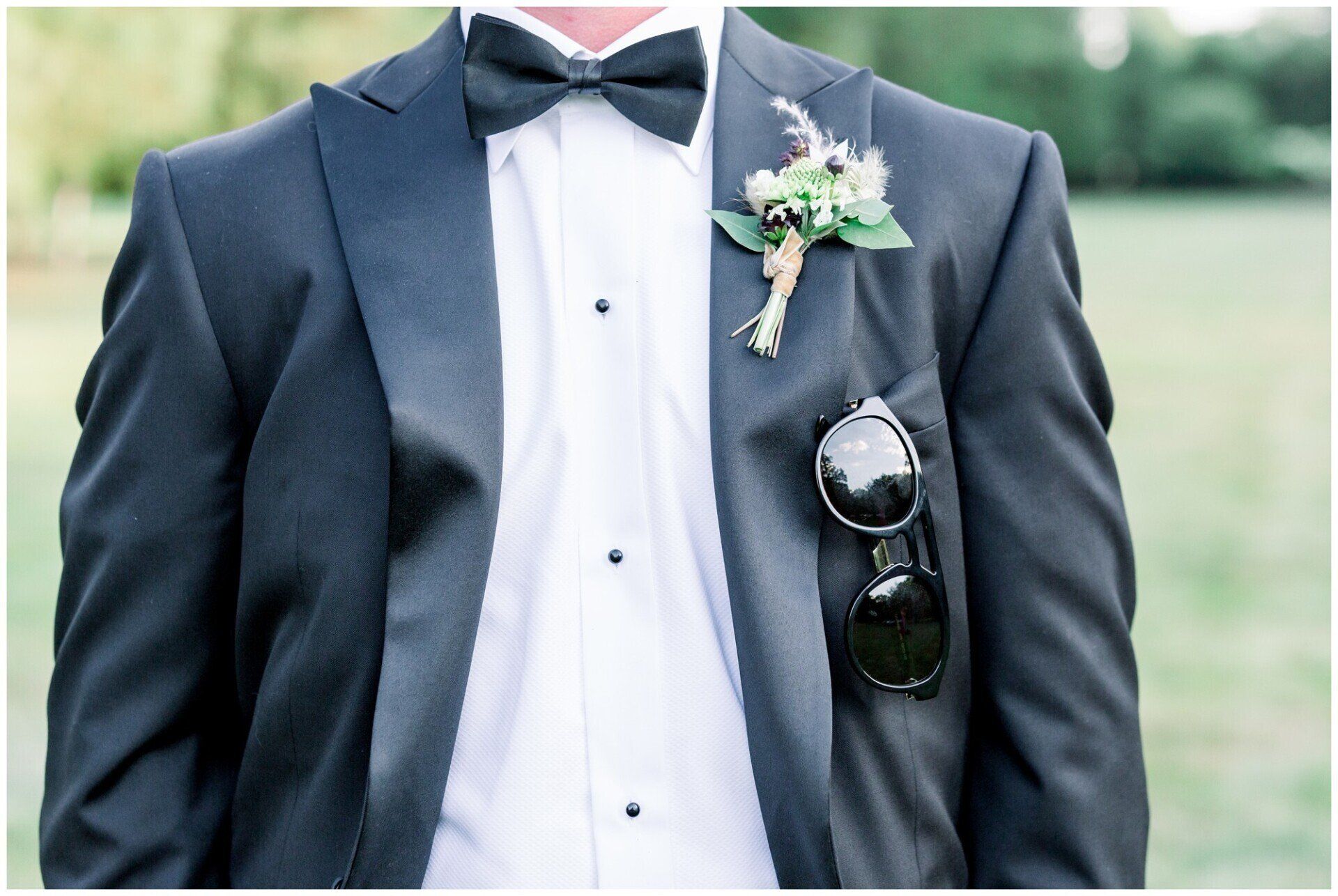 Man in dark suit with bow tie and boutonniere, sunglasses clipped to jacket, standing outdoors.