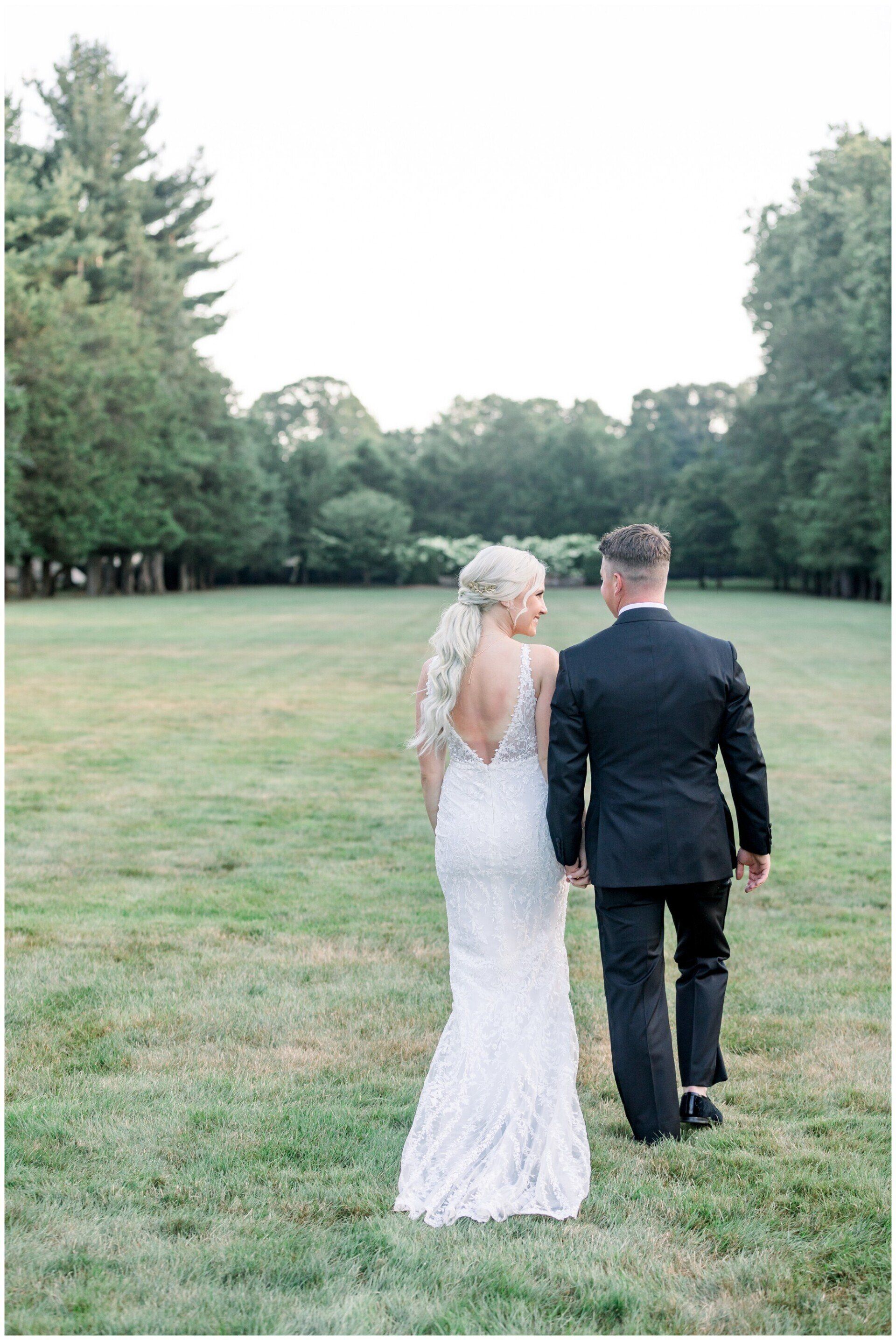 Couple in wedding attire walking hand in hand across a grassy field surrounded by trees