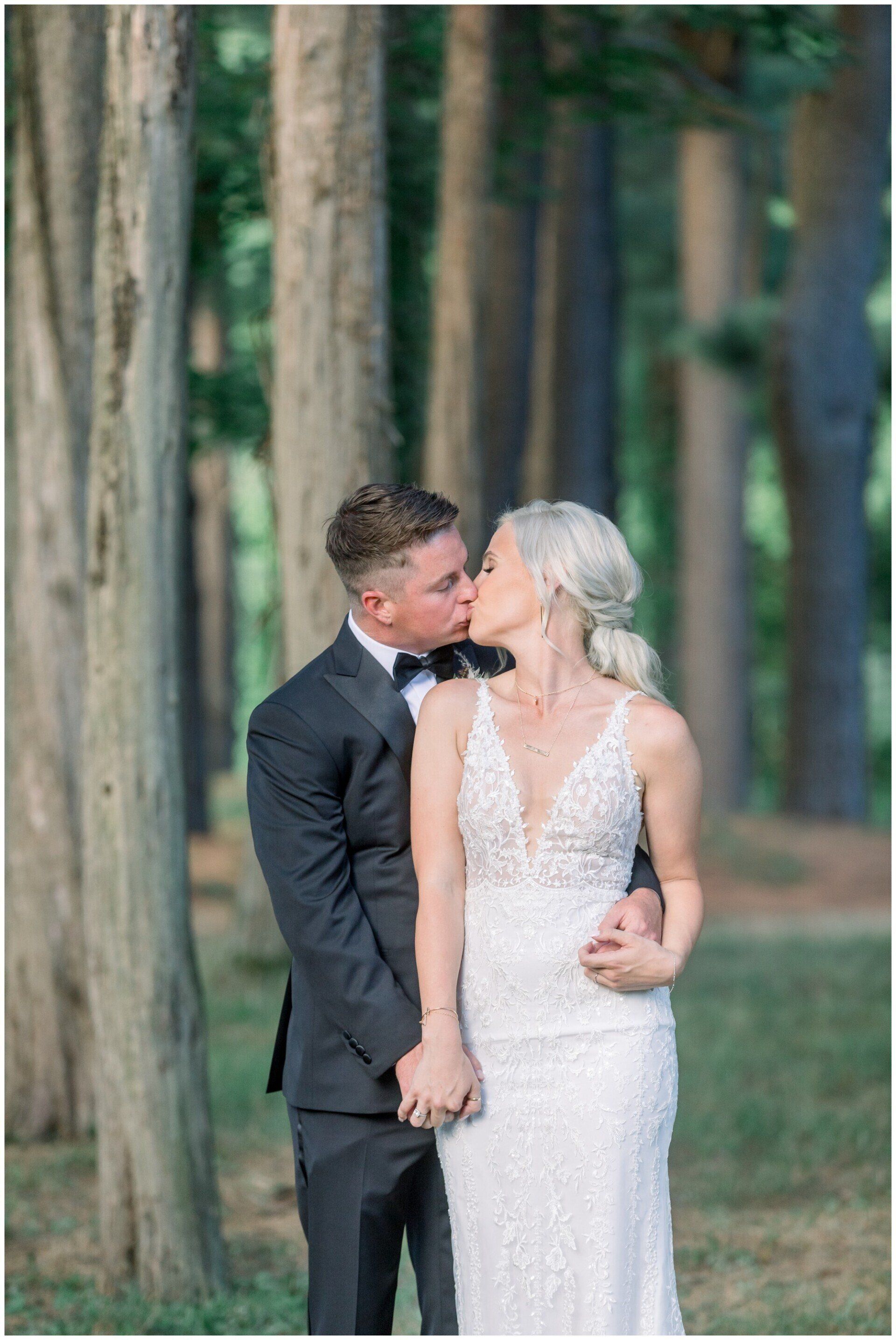 Two people in formal wear kissing in a wooded outdoor setting.
