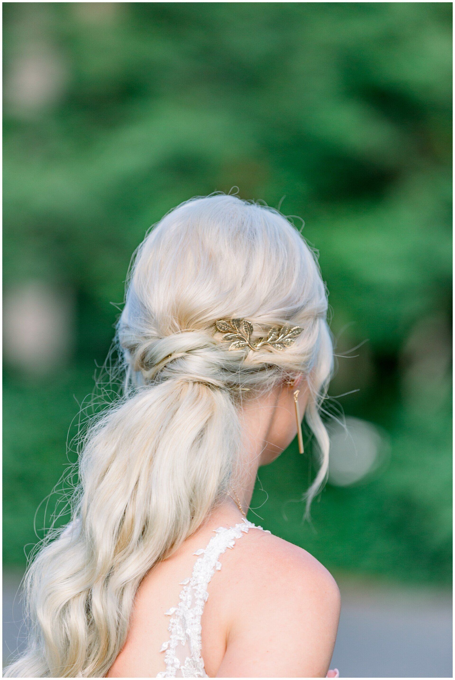 Blonde person seen from behind with a low ponytail, wearing a white lace dress against a green blurred background