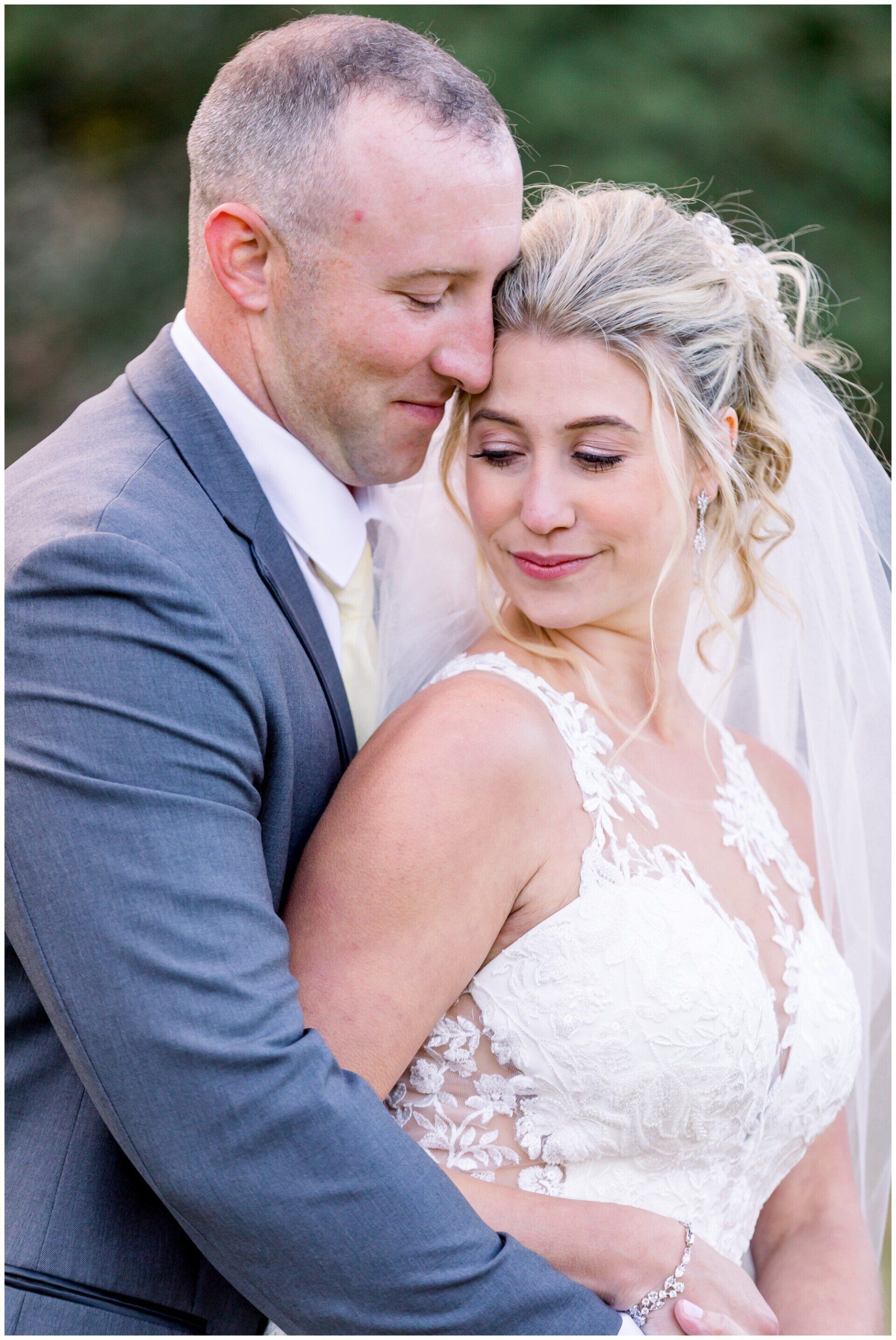 Bride and groom embracing outdoors, the bride in a white lace dress and veil, the groom in a gray suit
