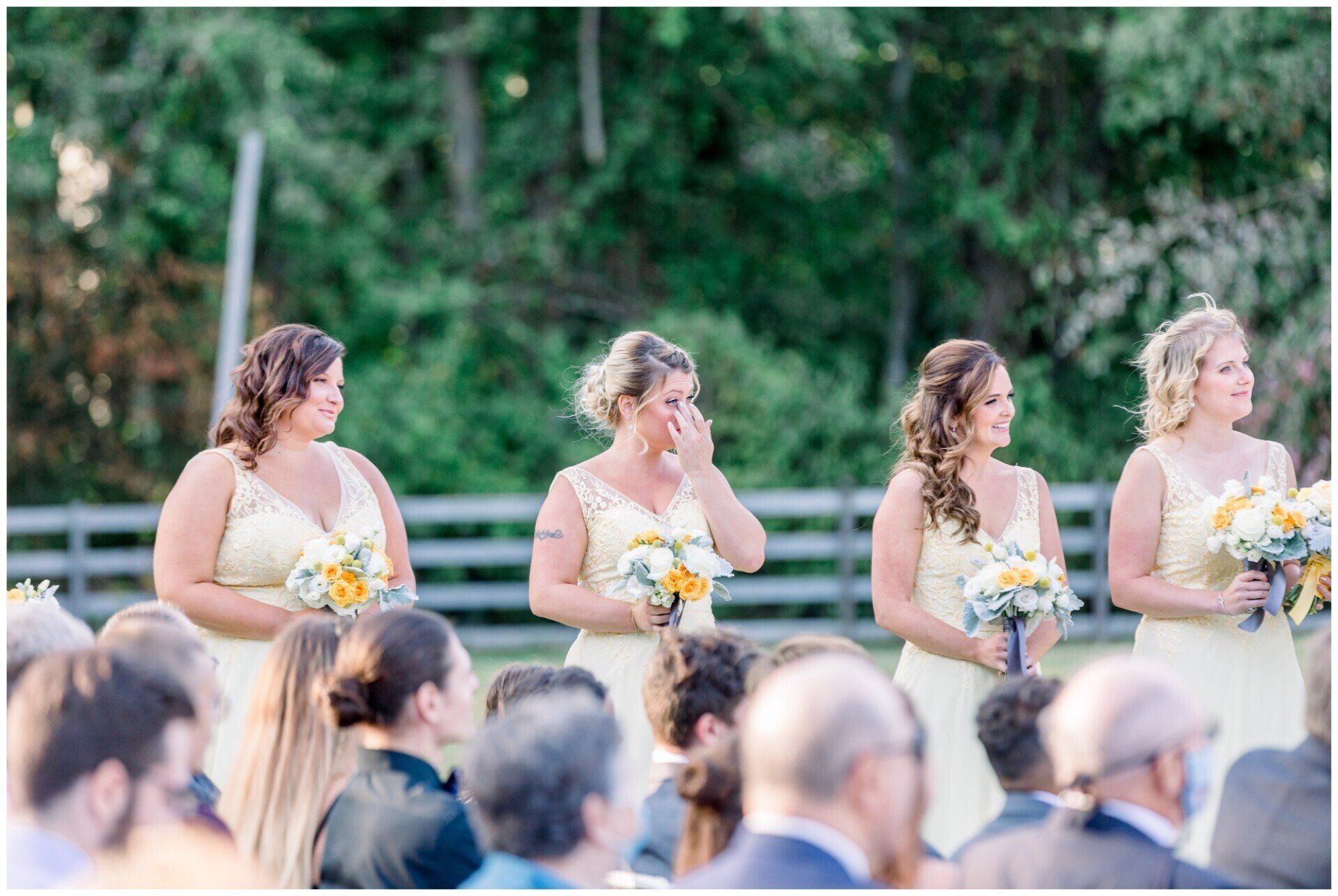 Bridesmaids in light dresses holding bouquets at an outdoor wedding ceremony, with guests seated in front.