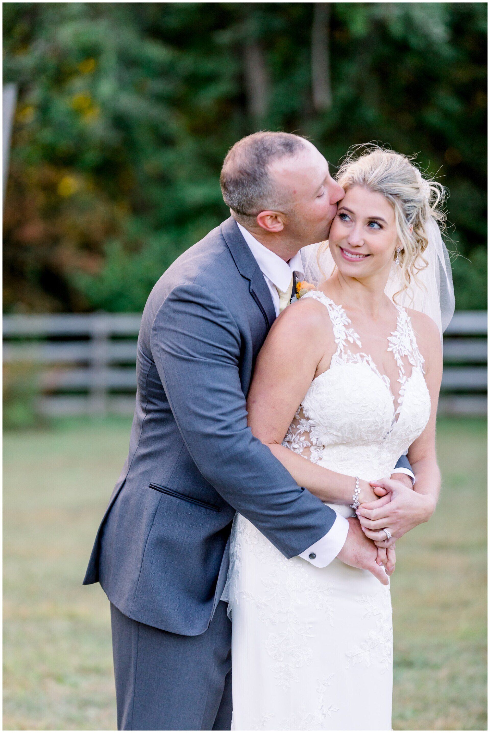 A groom kisses a smiling bride in a white dress outdoors.