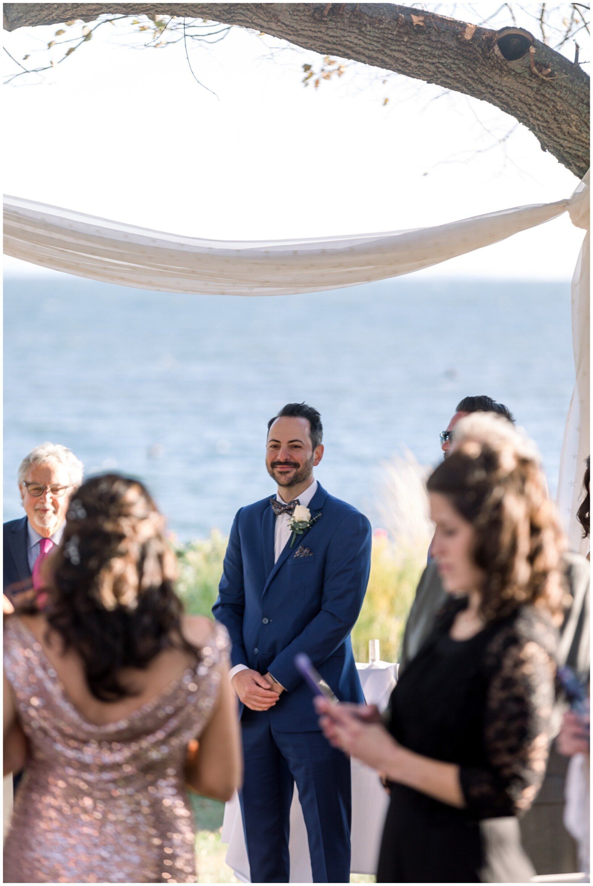 Man in blue suit standing at an outdoor wedding ceremony by the water