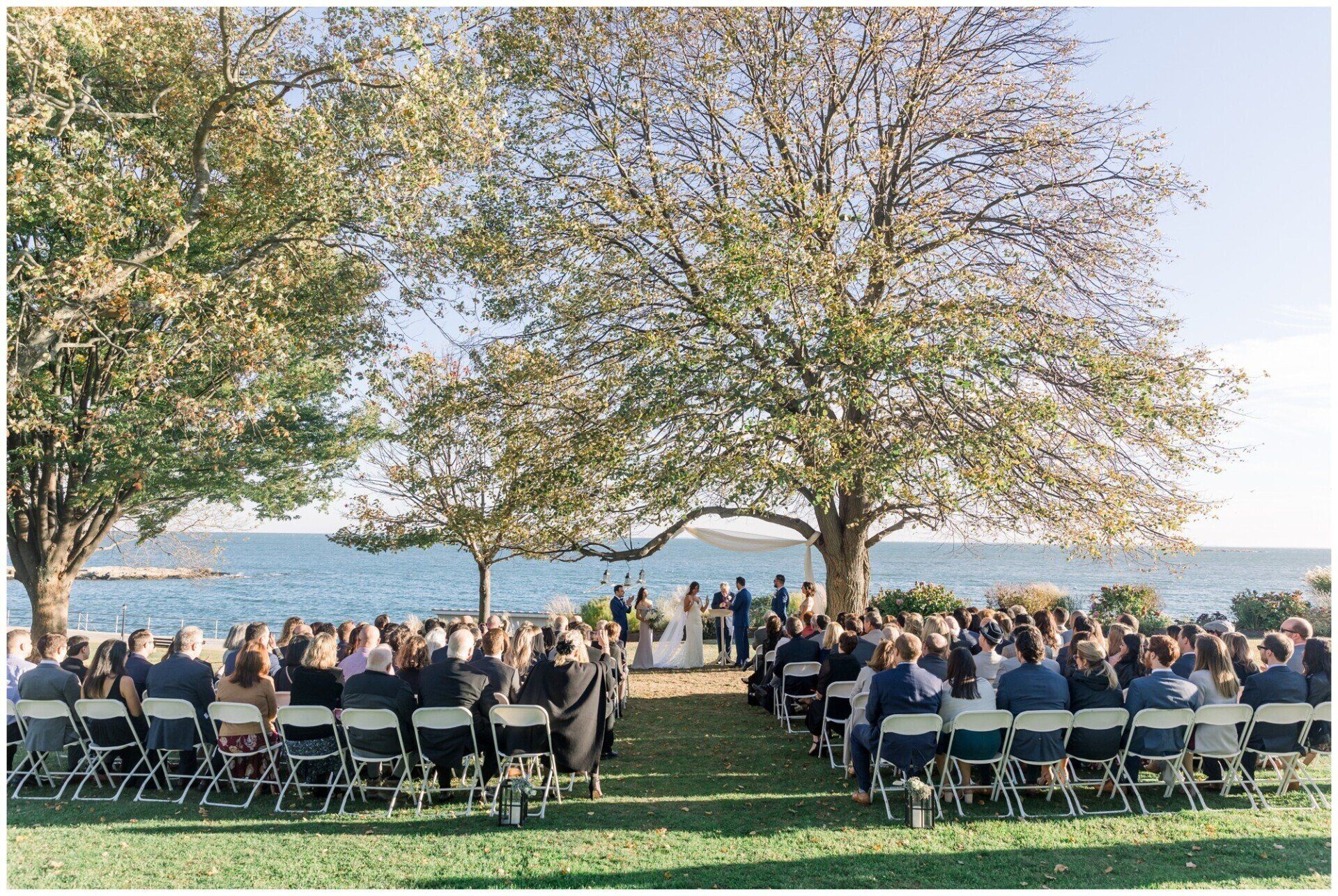 Outdoor wedding ceremony with guests seated on a lawn by the water under leafy trees