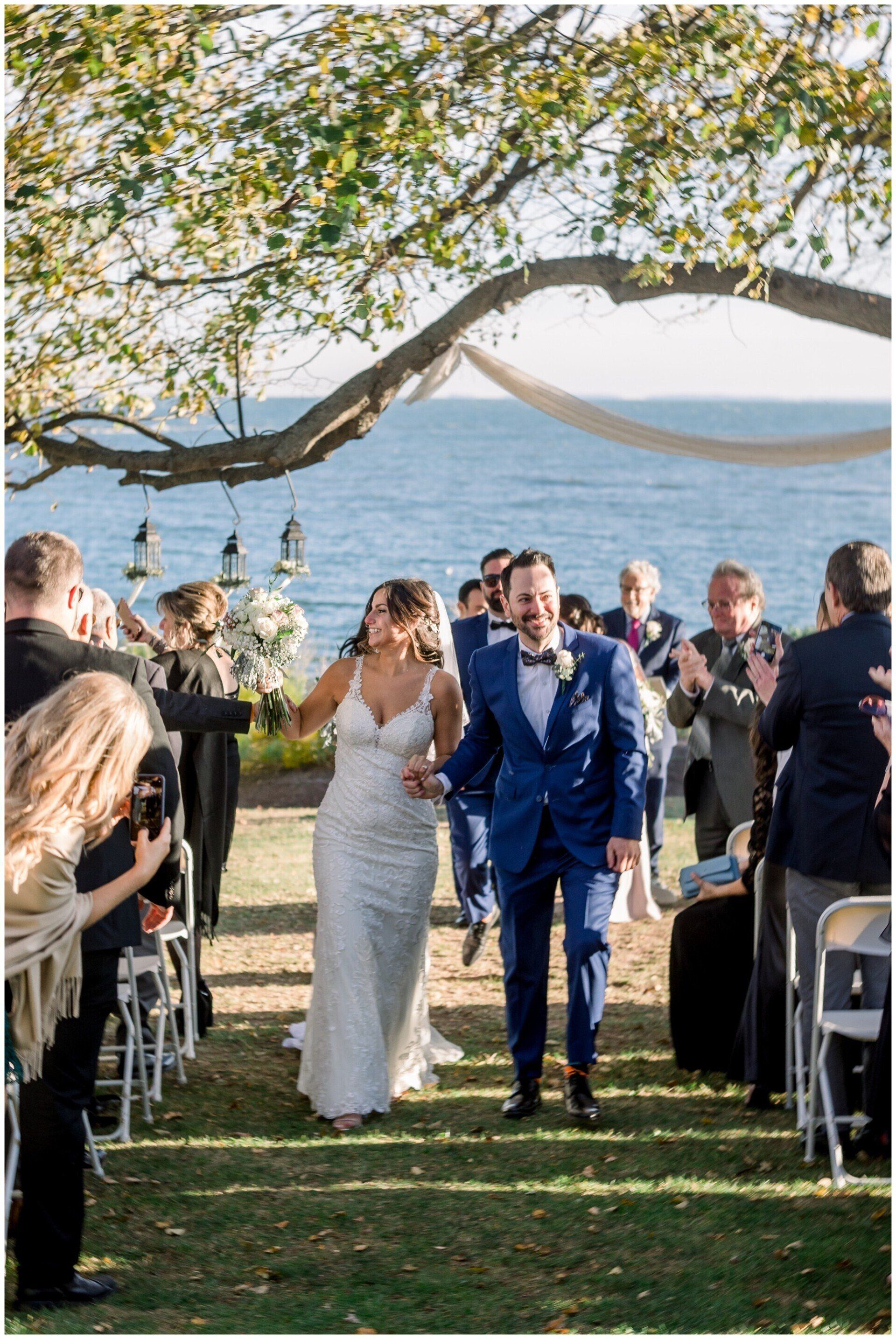Outdoor wedding ceremony with bride and groom walking down the aisle under a tree canopy by the water